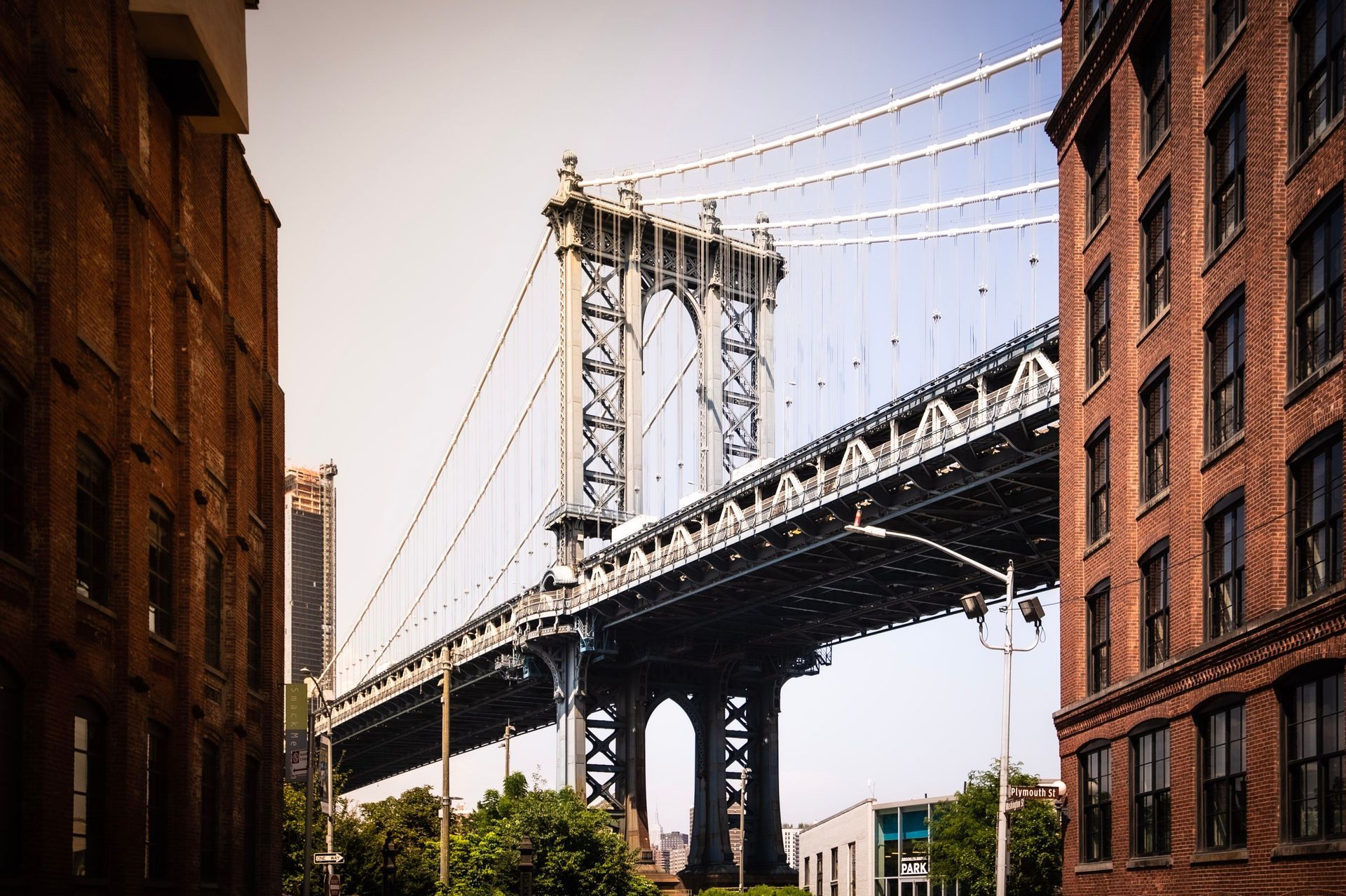 A steel suspension bridge rises between two multi-story brick buildings under a clear sky.