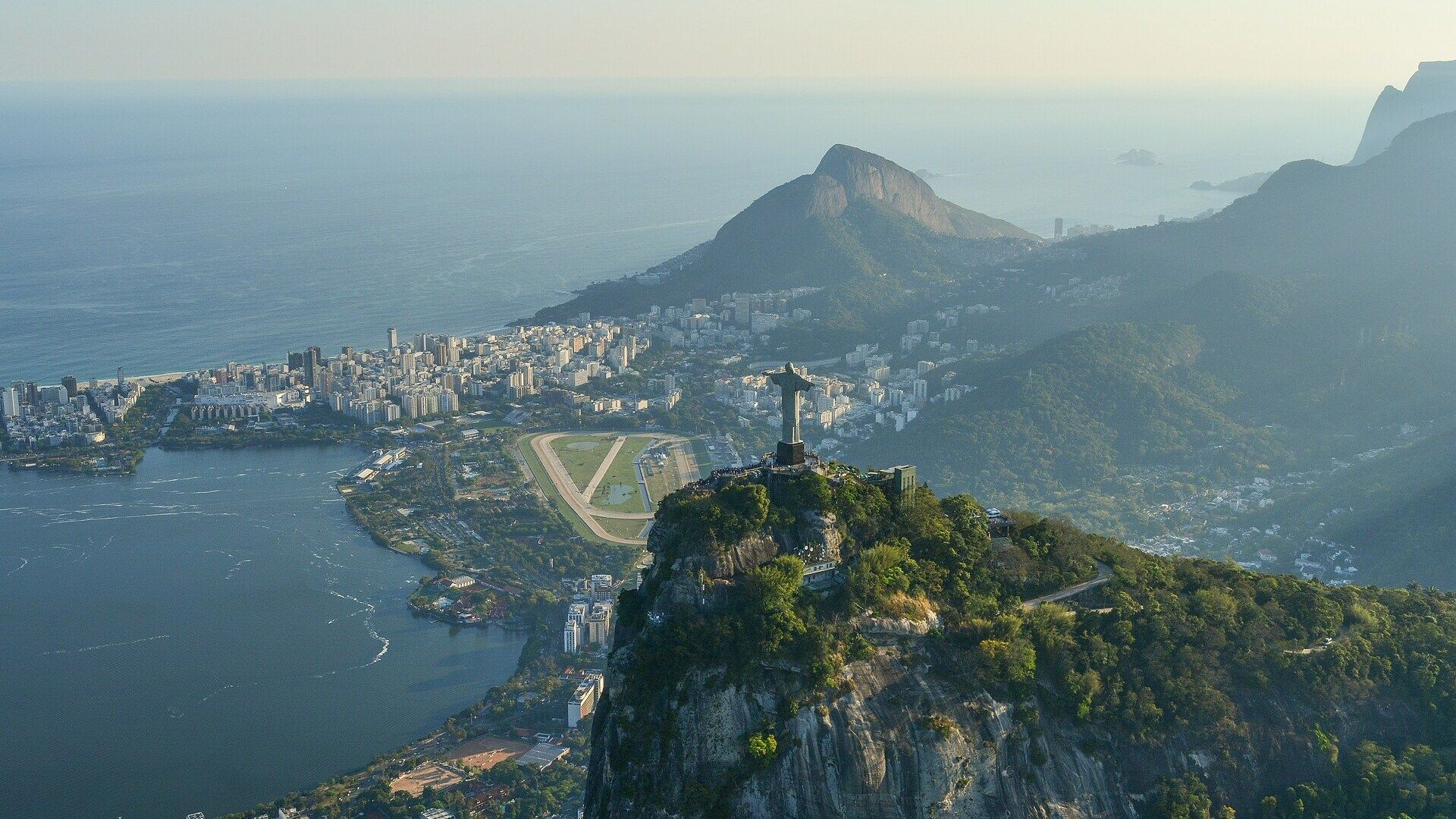 Una vista aerea della statua del Cristo Redentore su una montagna, che domina una città costiera con una grande laguna e l'oceano.