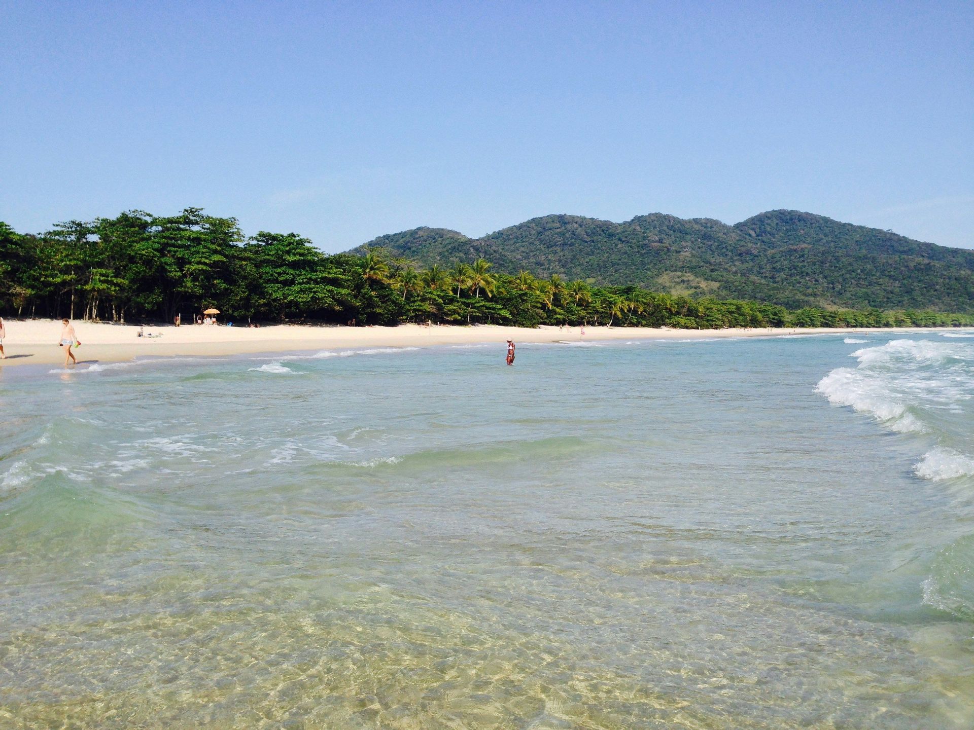 Acqua limpida dell'oceano lambisce una spiaggia di sabbia bianca, incorniciata da alberi verdi rigogliosi e montagne sotto un cielo blu.