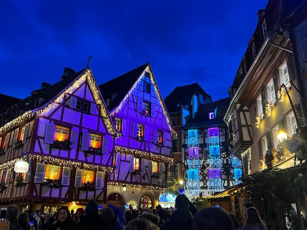 Edificios de entramado de madera iluminados con luces navideñas moradas y blancas sobre una calle concurrida por la noche