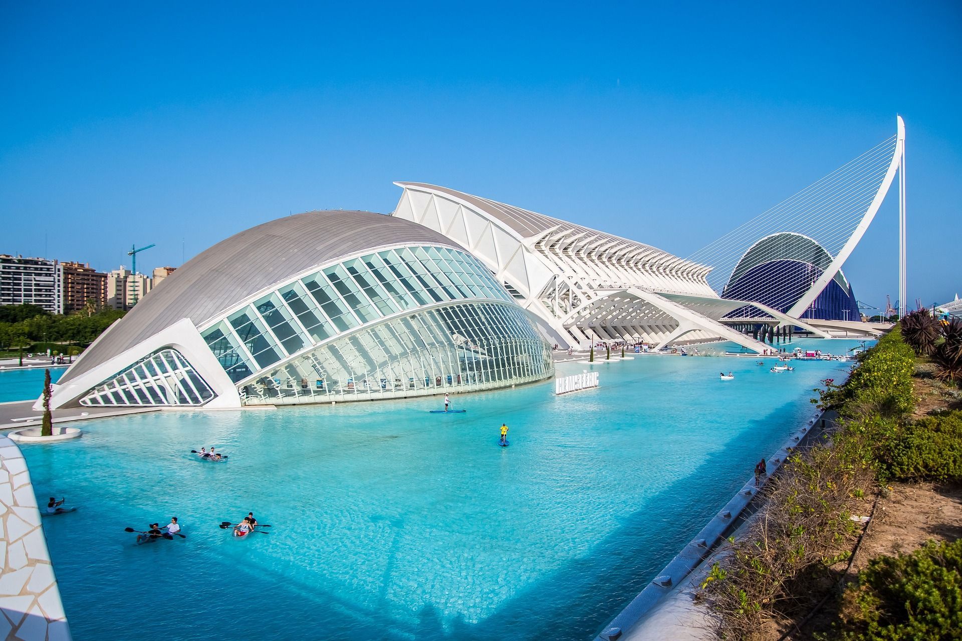 Moderni edifici architettonici bianchi vicino a una piscina di acqua turchese, con persone che praticano kayak e paddleboard sotto un cielo sereno.