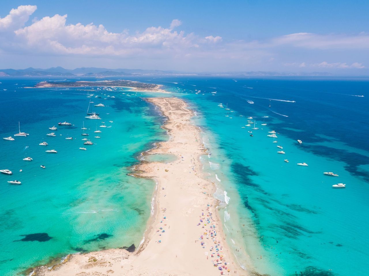 Una vista aerea di un lungo istmo sabbioso circondato da un mare turchese, con numerose barche e yacht ancorati su entrambi i lati sotto un cielo blu.