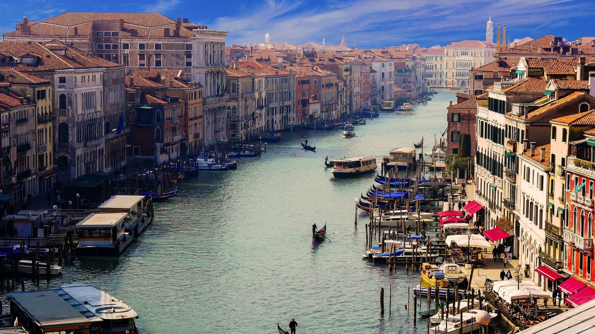 Una vista dall'alto di un ampio canale pieno di barche e gondole, fiancheggiato da edifici storici sotto un cielo blu.