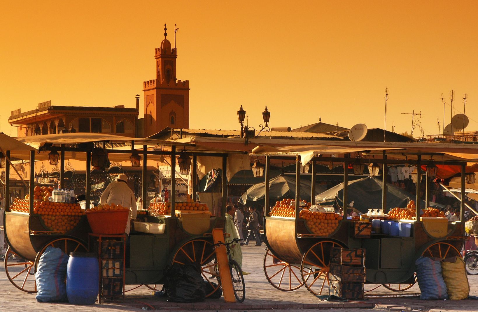Two wooden carts filled with oranges stand in a bustling market square under an orange sky, with a minaret in the background.