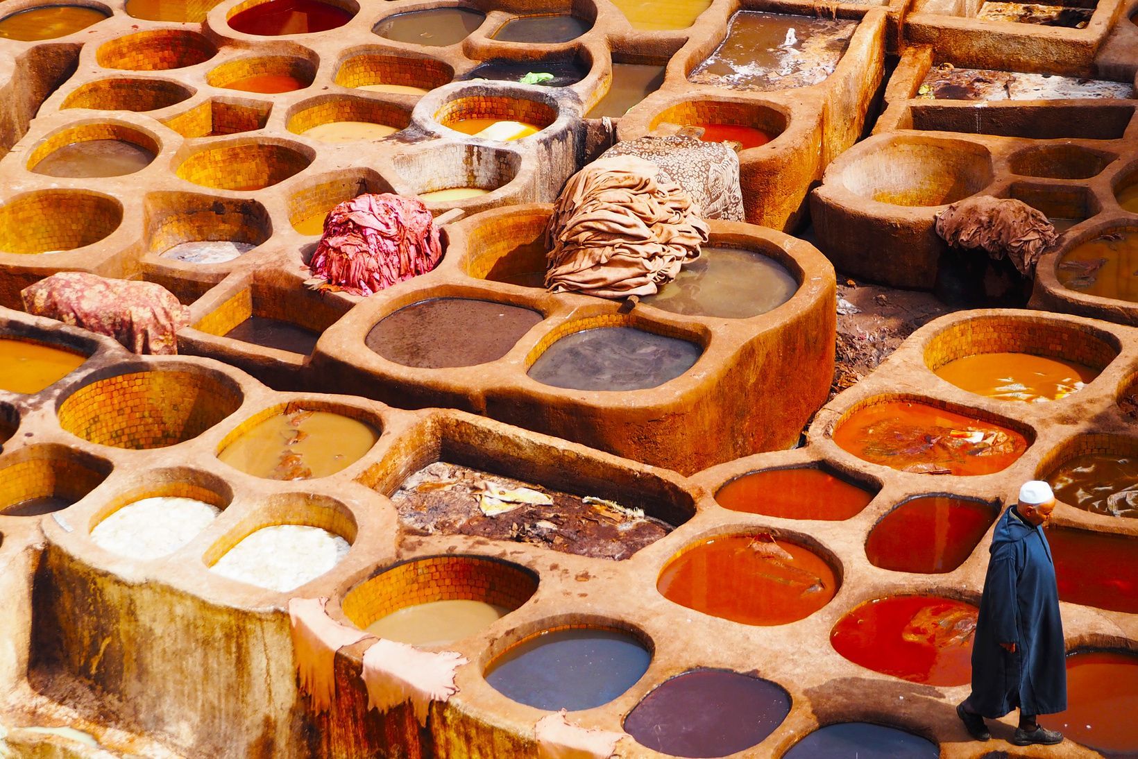 A man in a blue djellaba walks past large, stone vats filled with colorful dyes at an outdoor tannery.