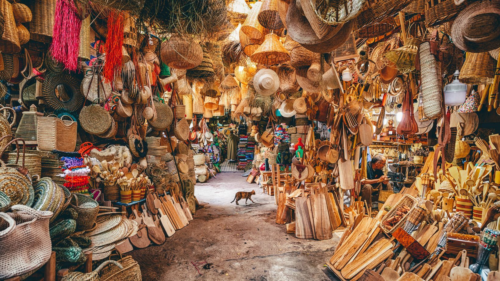 A cat walks down the aisle of a narrow market stall crowded with woven baskets, straw hats, and wooden crafts.