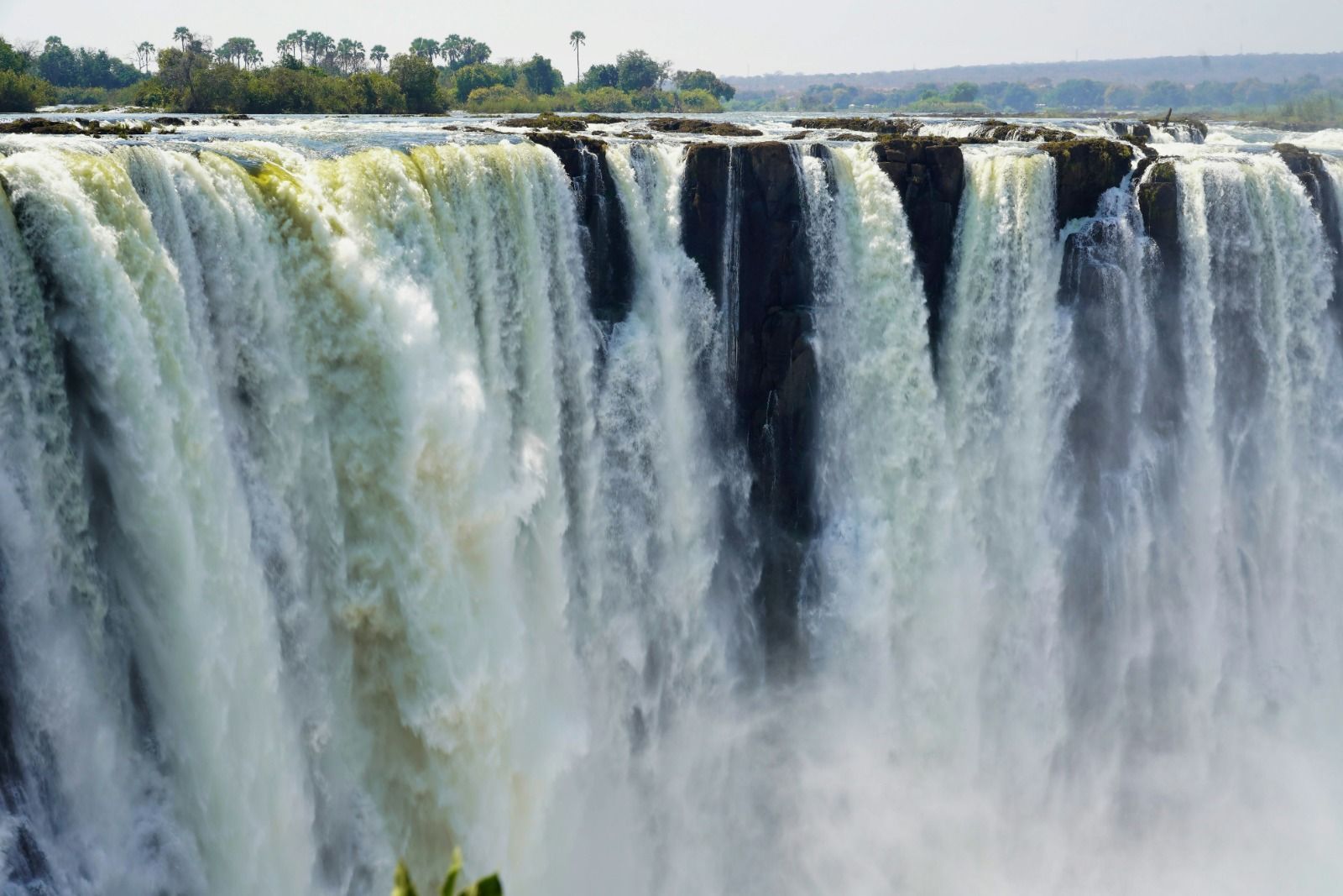 Una cascata ampia e potente precipita da un dirupo roccioso, generando una grande nuvola di nebbia alla sua base con alberi in lontananza.