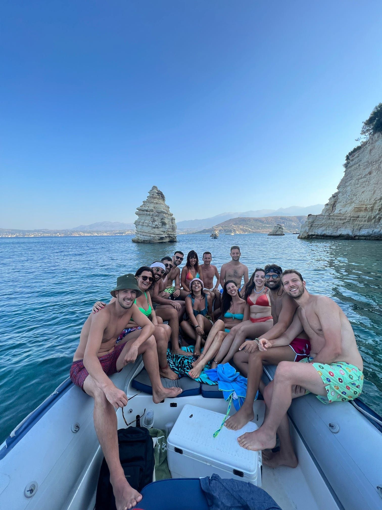 Un grupo sonriente de WeRoad en traje de baño posa en un barco en el mar, con grandes formaciones rocosas y acantilados costeros detrás.