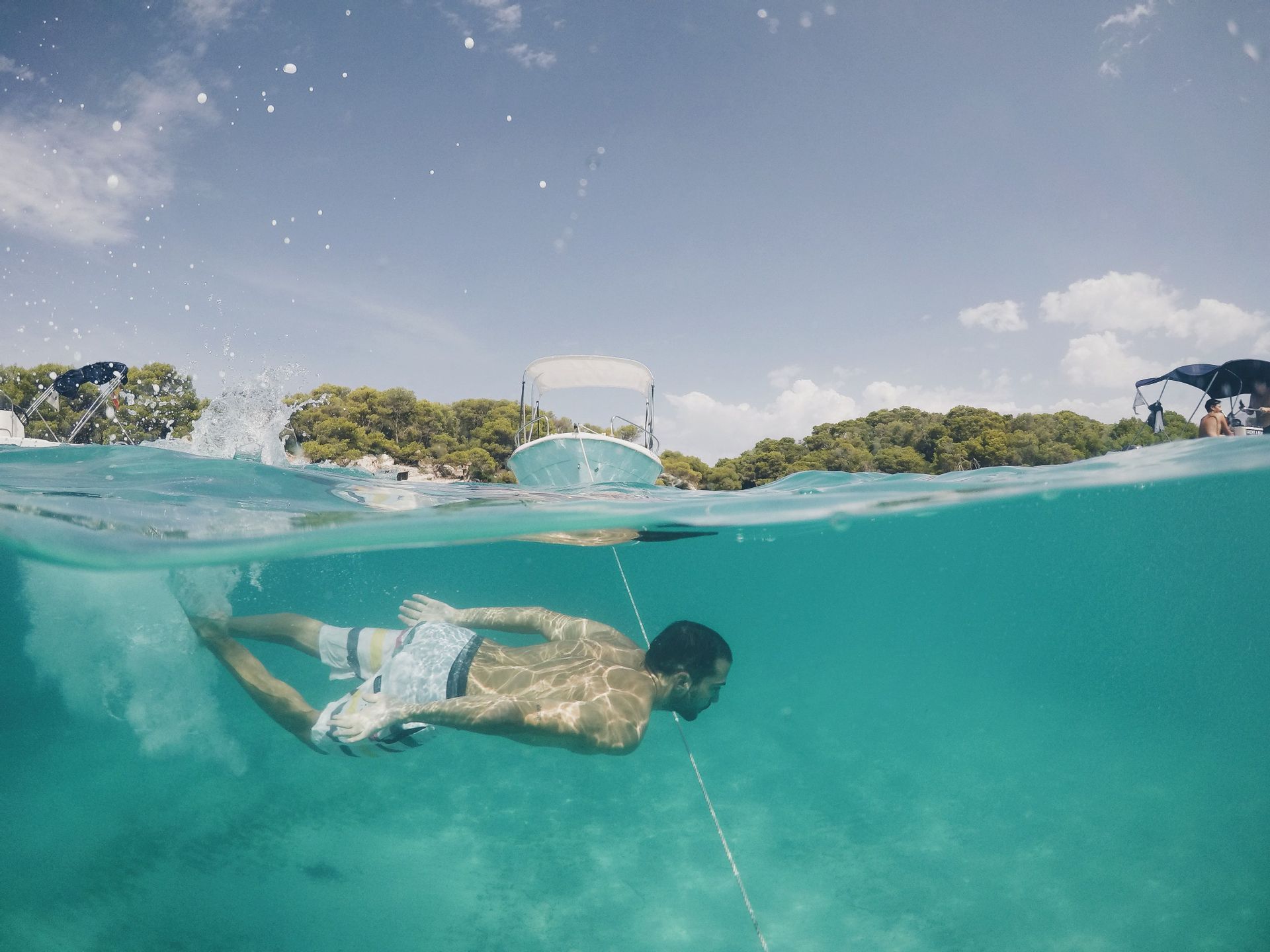 Vue aquatique et aérienne d'un homme nageant sous l'eau dans une mer turquoise et limpide, avec des bateaux et une côte arborée visibles en surface.