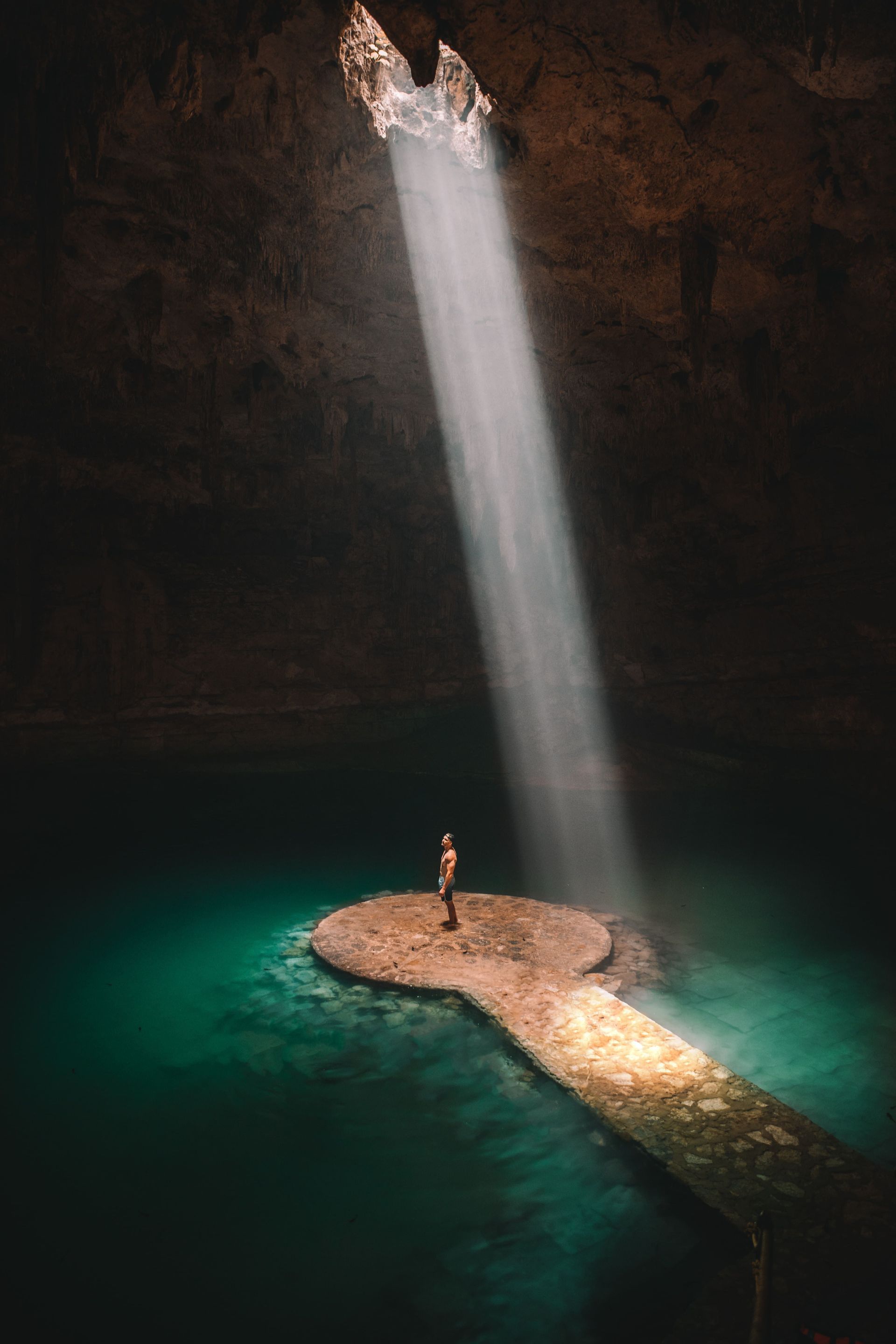 Una persona si trova su una piattaforma di pietra in un cenote, illuminata da un raggio di luce proveniente da un'apertura nel soffitto della grotta.