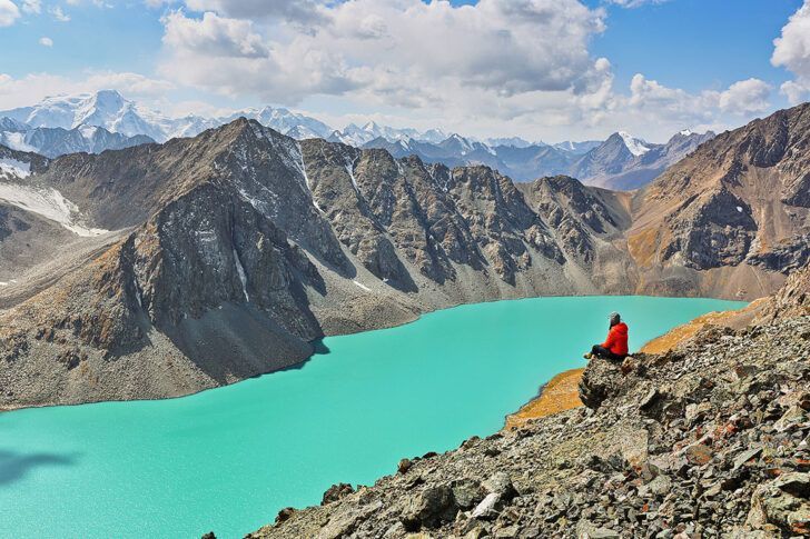 Une personne en veste rouge assise sur une falaise rocheuse surplombant un lac alpin turquoise vif entouré de montagnes sous un ciel nuageux.