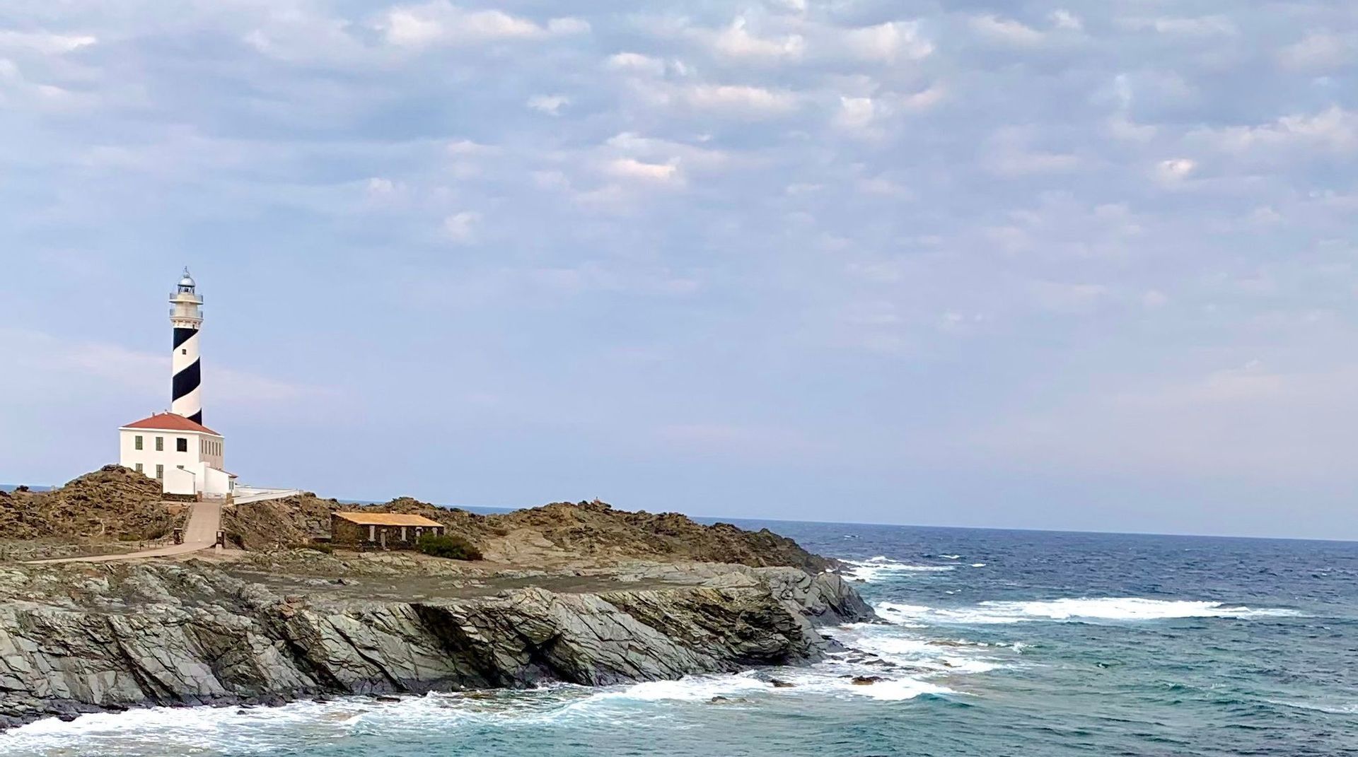 Ein schwarz-weiß spiralförmiger Leuchtturm steht auf einer felsigen Klippe mit Blick auf das Meer unter einem bewölkten Himmel.