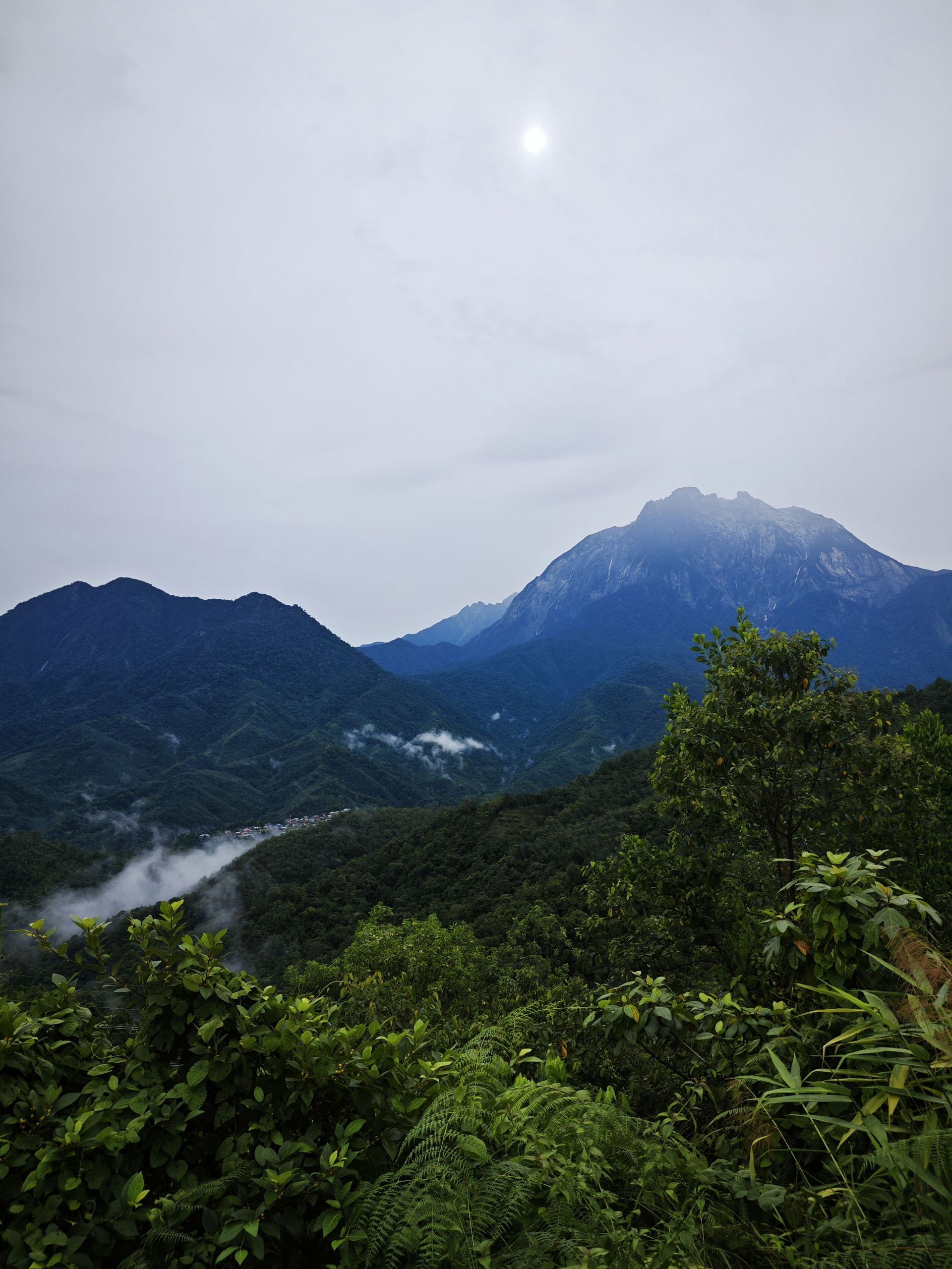 Vista de un exuberante valle de selva verde con una gran montaña a la distancia bajo un cielo brumoso y el sol visible.