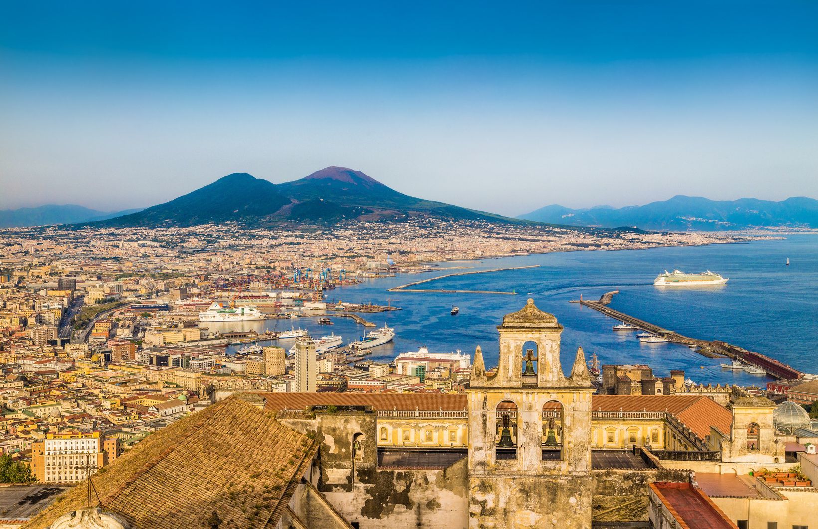 Un campanile in pietra si affaccia su una vasta città costiera e su una grande baia, con un prominente vulcano visibile sullo sfondo sotto un cielo azzurro e limpido.