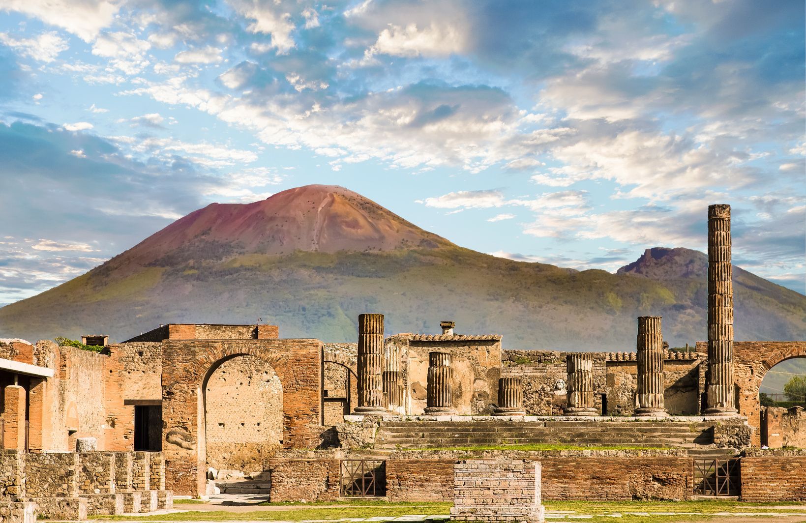 Ruinas antiguas con columnas y arcos de piedra se alzan ante un gran volcán bajo un cielo parcialmente nublado.