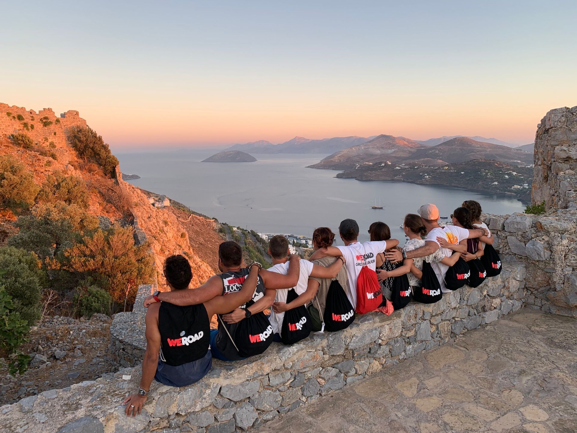 Un gruppo WeRoad siede abbracciato su un muretto di pietra, ammirando il mare con isole al tramonto.