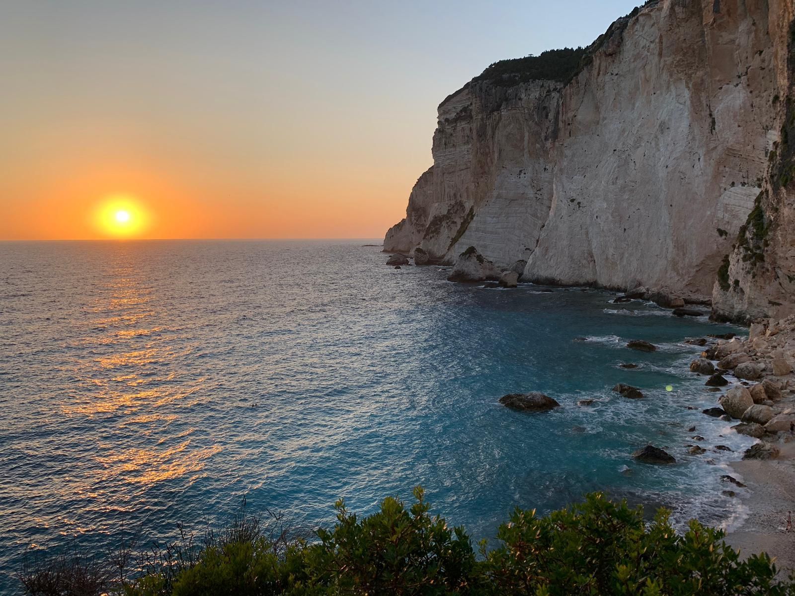 The sun sets over a calm ocean next to towering white limestone cliffs under a clear sky.
