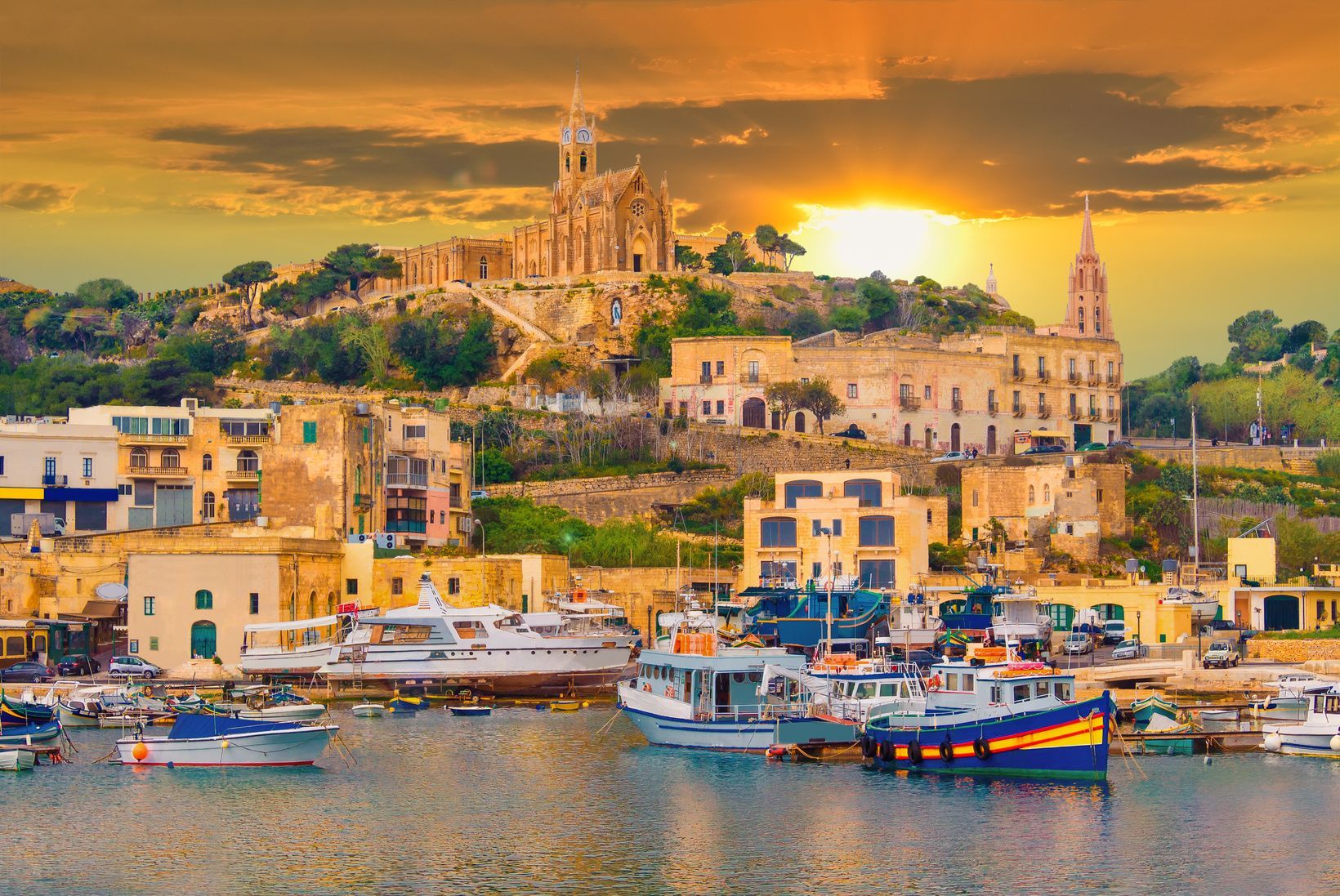 A harbor filled with boats and yachts in front of a hillside town featuring a large church under a golden sunset.