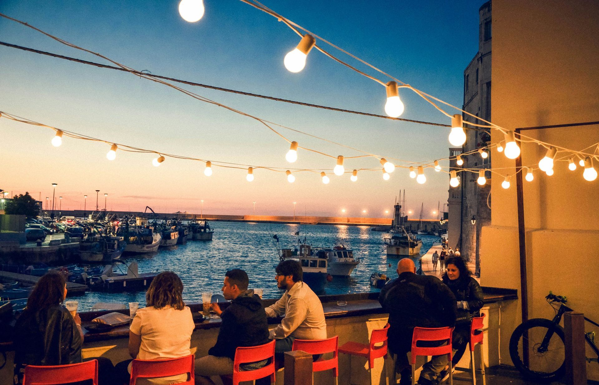 Un viaggio di gruppo WeRoad si gode un drink in un bar all'aperto con vista su un porto turistico al tramonto sotto le luci a festa.