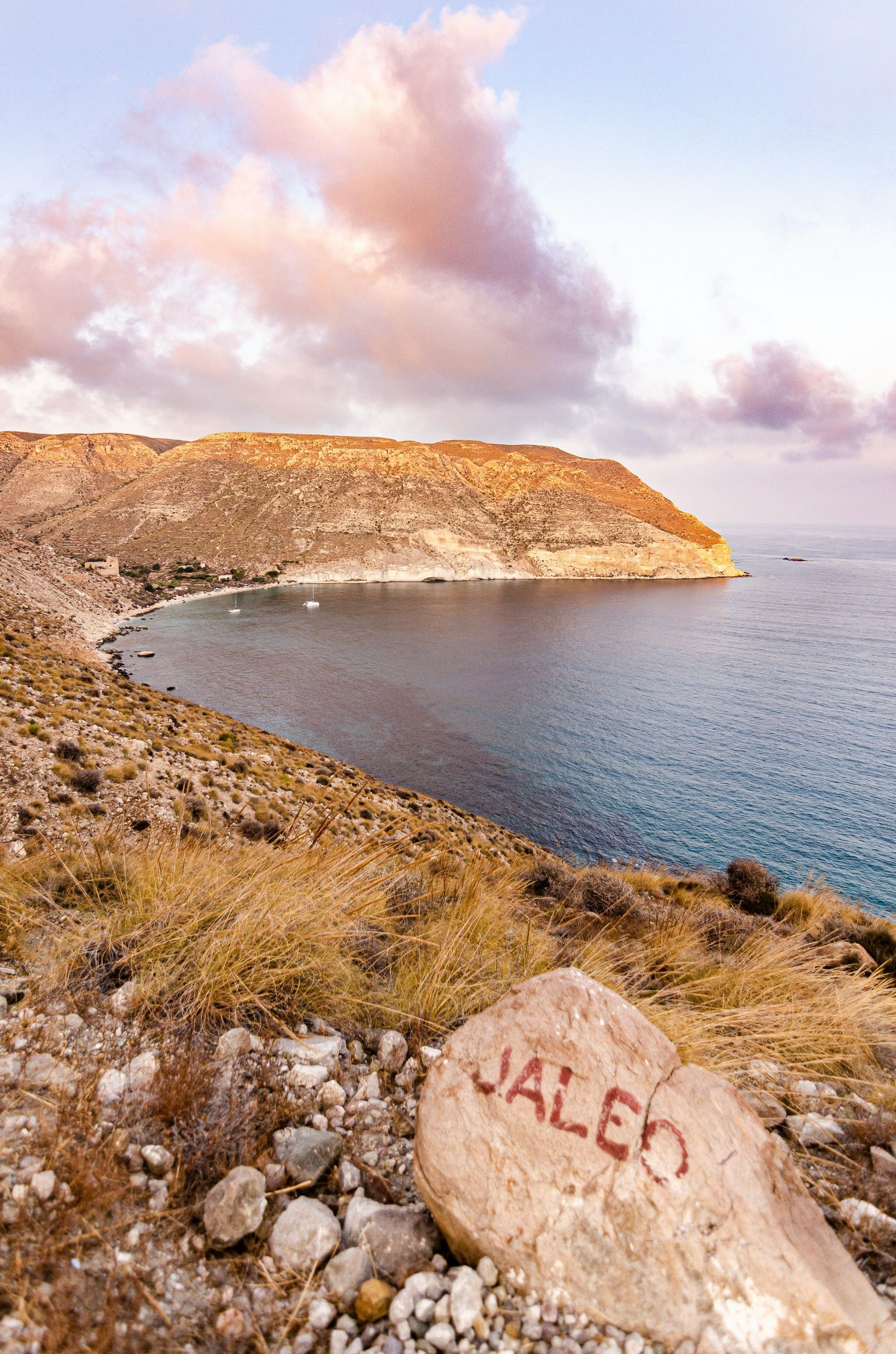 Una roca pintada con la palabra 'JALEO' se asienta en una colina seca y cubierta de hierba, con vistas a una cala con veleros y acantilados áridos al atardecer.