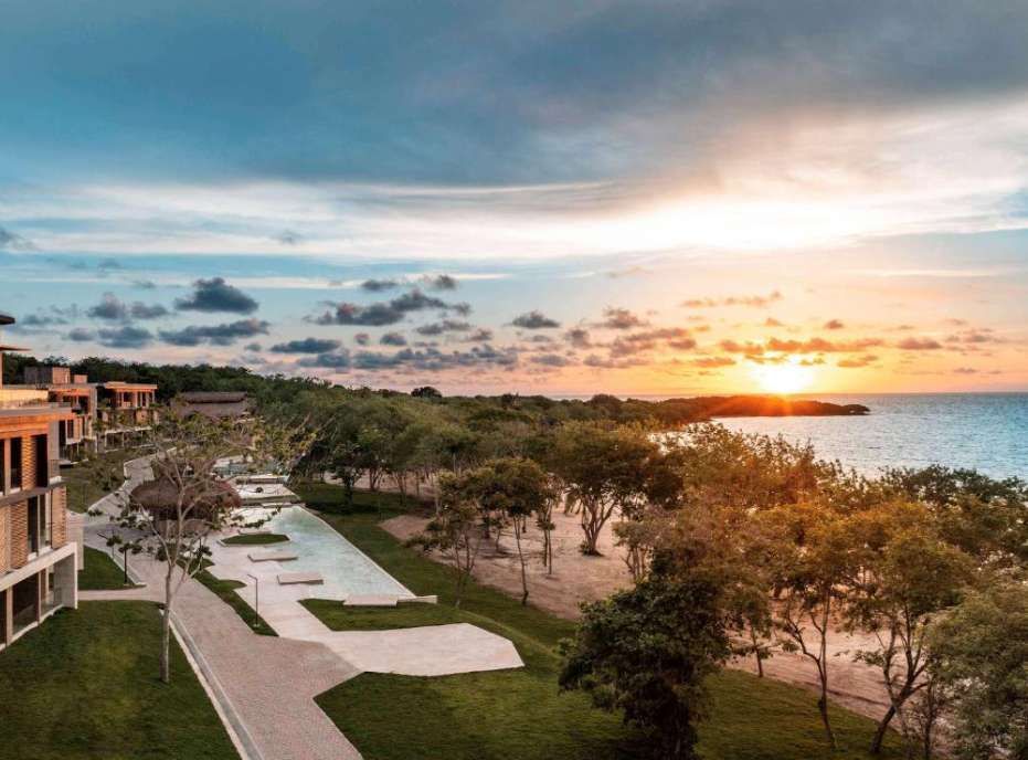 An aerial view of a coastal resort with modern buildings, pools, and trees overlooking the ocean at sunset.