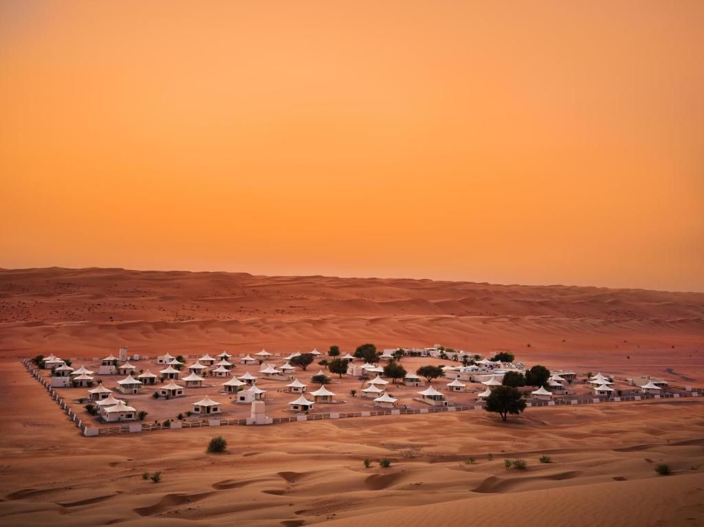Un campo tendato bianco si trova in un recinto tra dune di sabbia ondulate sotto un cielo arancione.