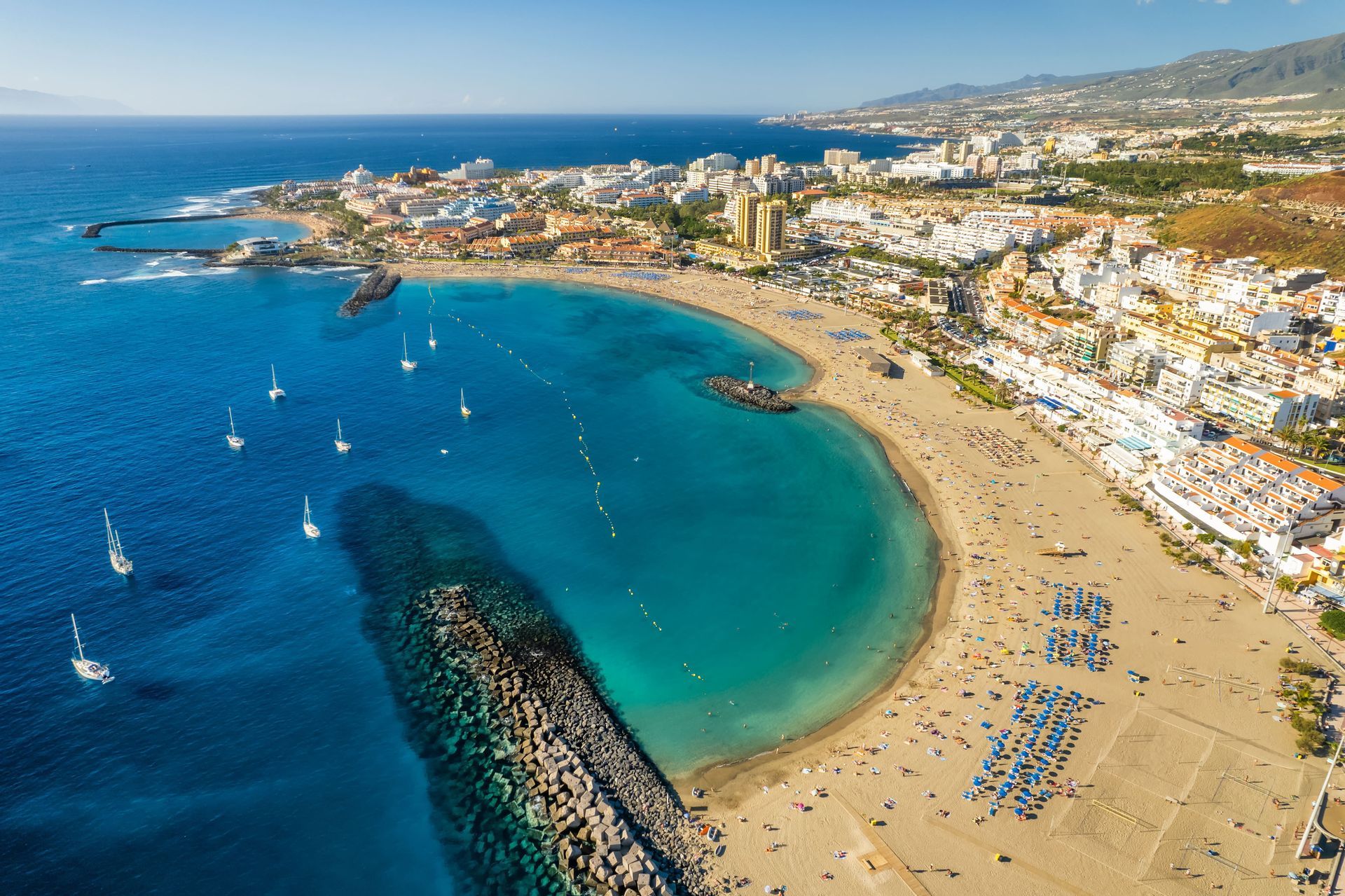Luftaufnahme einer Küstenstadt mit einem belebten Sandstrand an einer türkisfarbenen Bucht voller Segelboote unter klarem blauem Himmel.