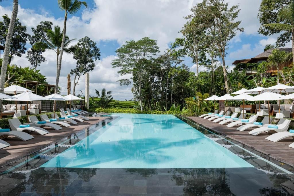 Ein langer Swimmingpool in einem tropischen Resort, gesäumt von Reihen weißer Liegen und Sonnenschirmen unter einem teilweise bewölkten Himmel.