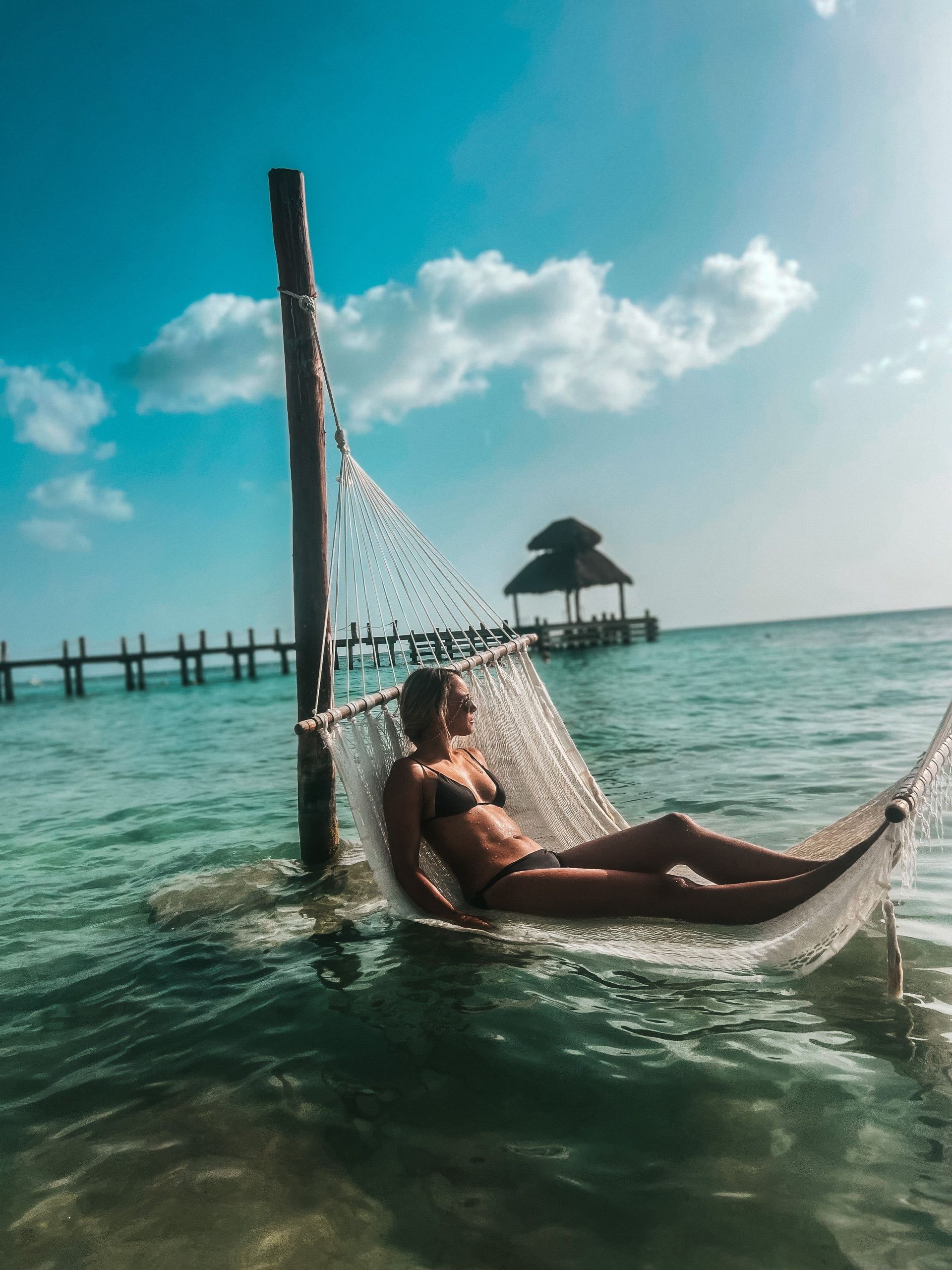 Una donna in bikini nero si rilassa su un'amaca bianca sospesa sull'acqua turchese dell'oceano, con un molo sullo sfondo.