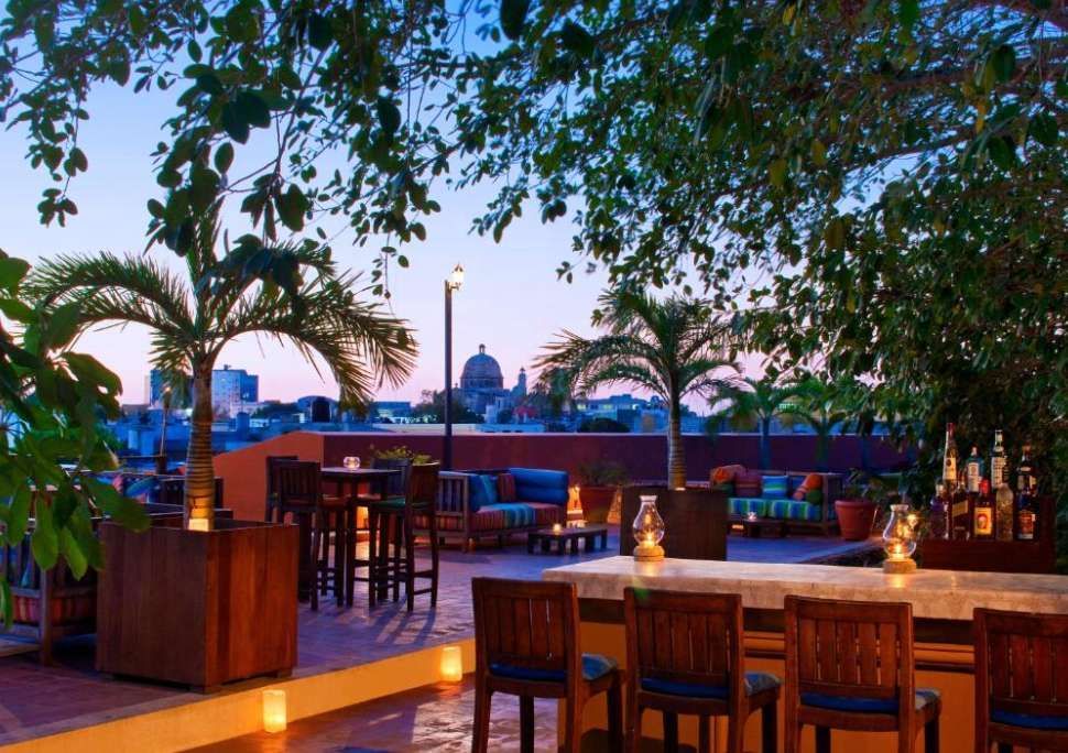 Un bar panoramico sul tetto, con mobili in legno e palme, si affaccia sullo skyline di una città con un edificio a cupola al tramonto.