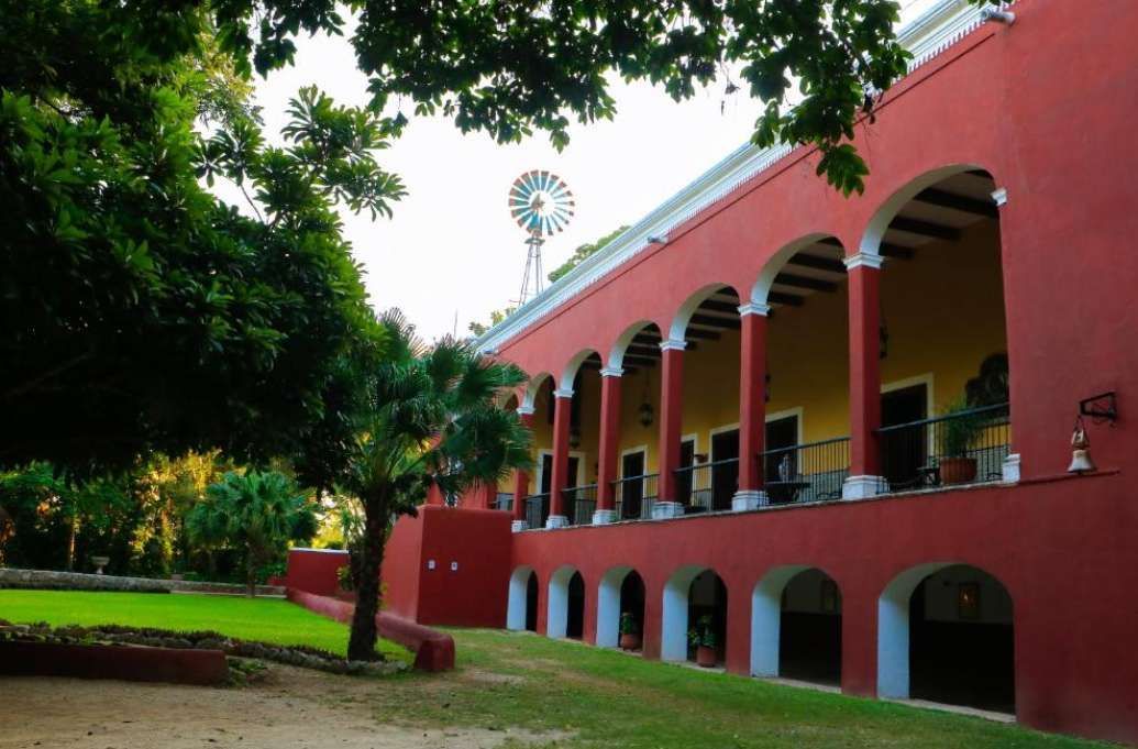 Una hacienda rossa a due piani con una veranda ad arco e un mulino a vento sul tetto si erge accanto a un prato rigoglioso con palme.