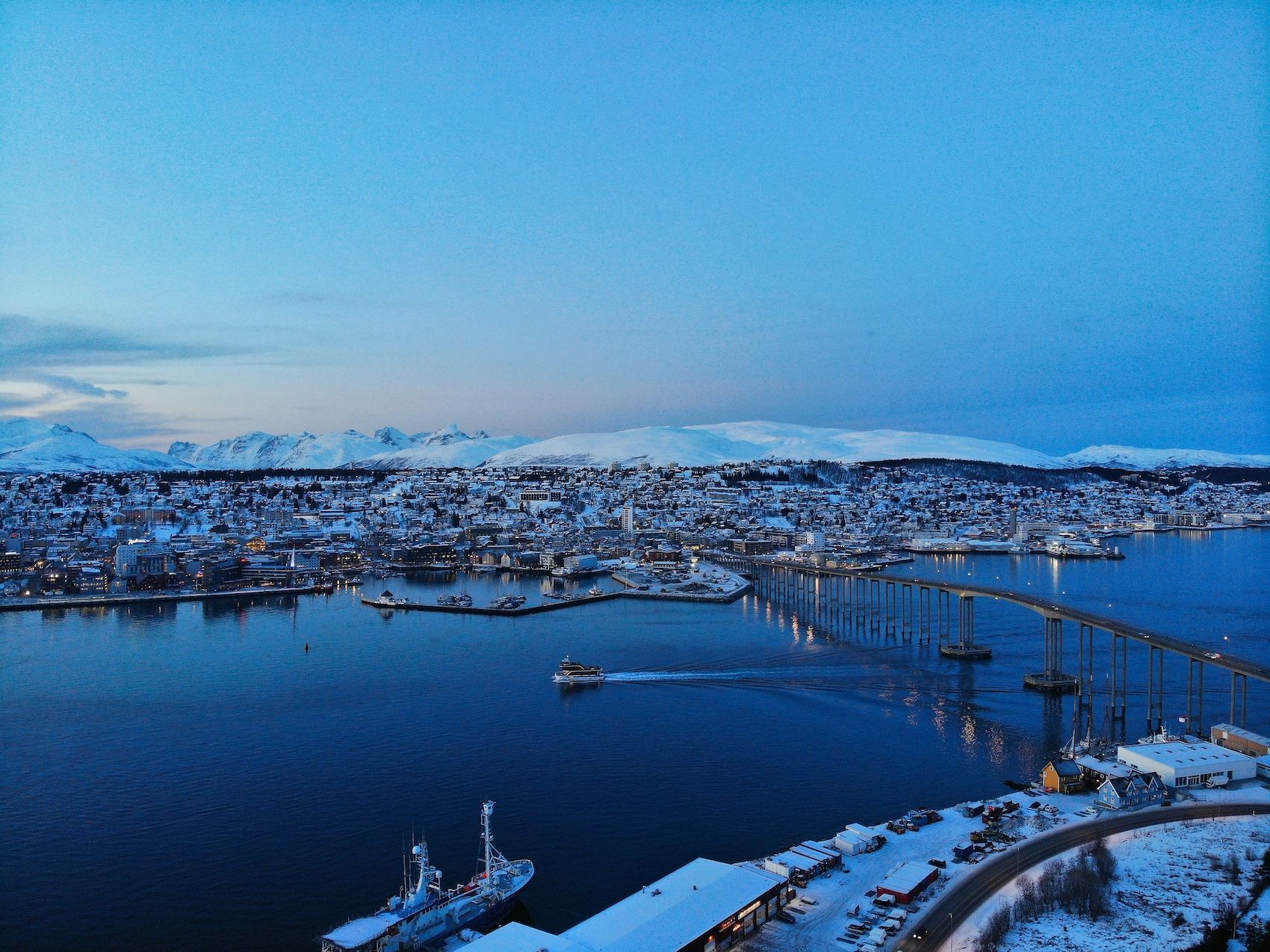 Une vue aérienne d'une ville côtière enneigée au crépuscule, avec un long pont sur l'eau et des montagnes en arrière-plan.