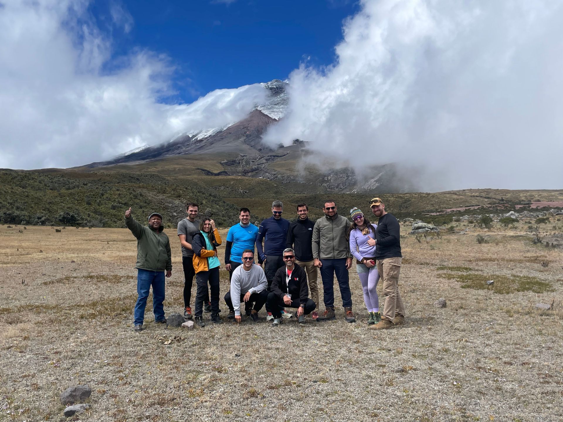 A WeRoad group trip poses for a photo in a mountain landscape with a large, snow-capped peak partially covered by clouds.