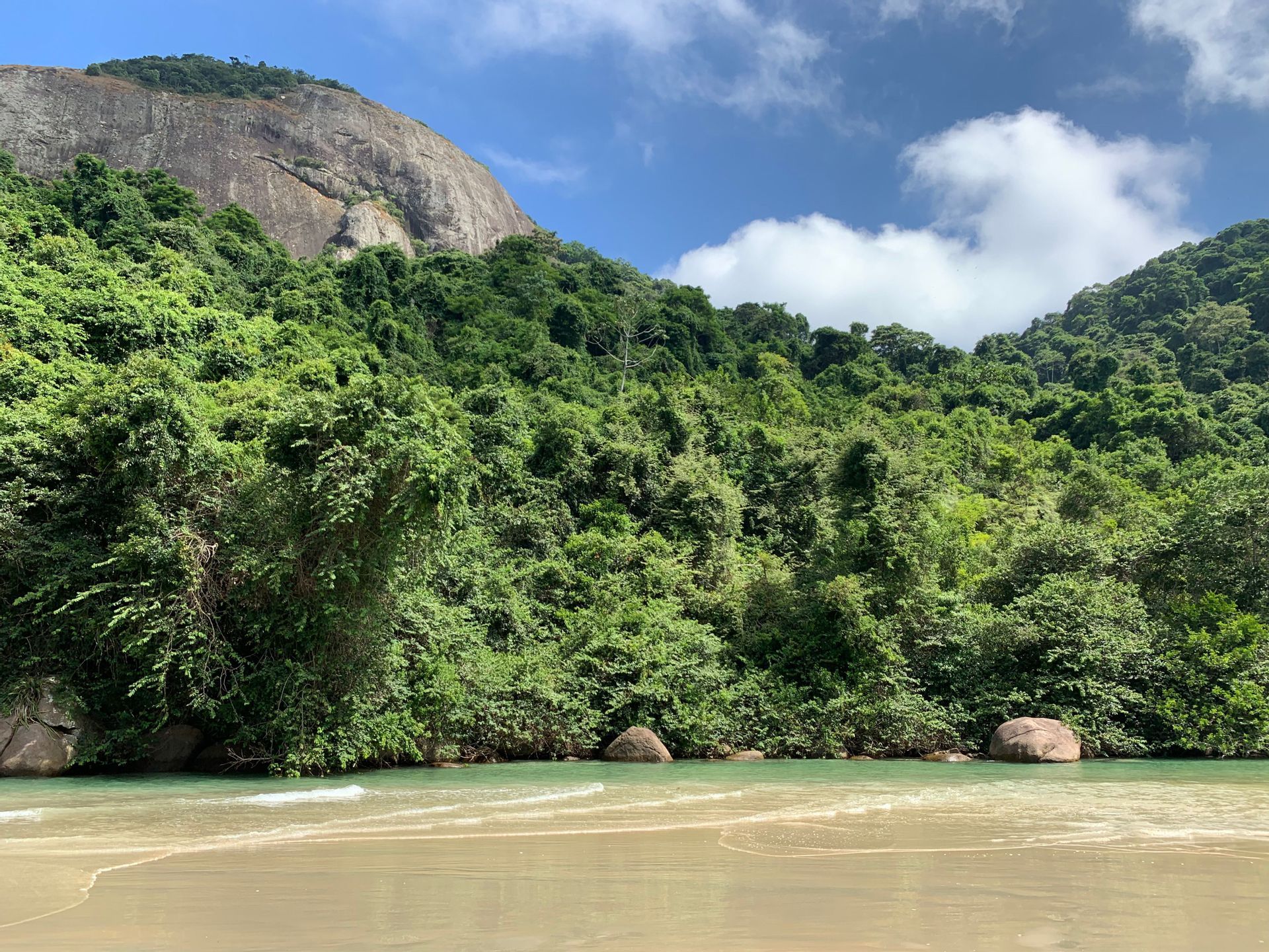 Una spiaggia sabbiosa con acqua calma e verde chiaro, ai piedi di una montagna lussureggiante ricoperta di giungla, sotto un cielo azzurro.