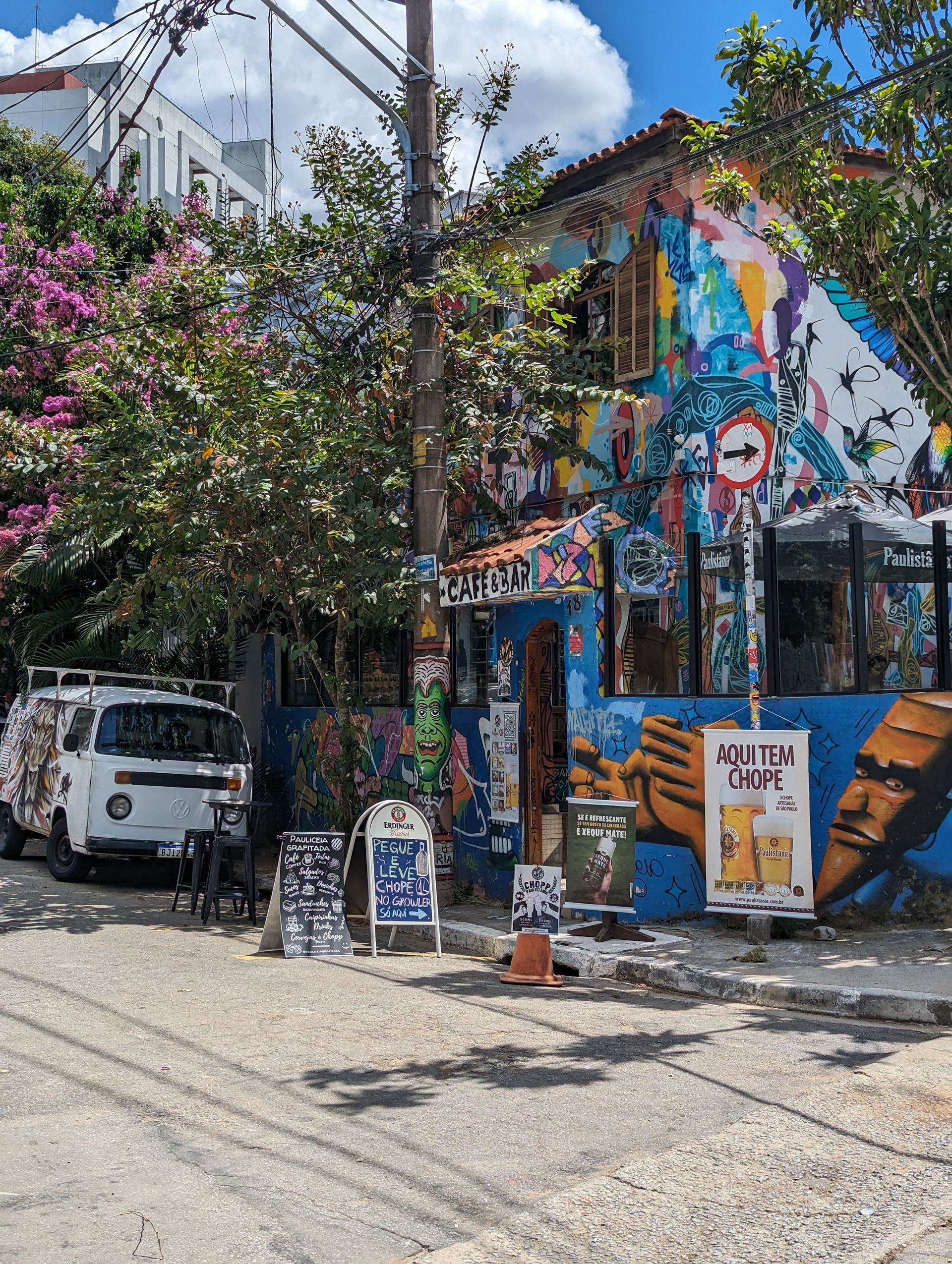 Un café y bar de esquina en una calle de la ciudad, sus paredes cubiertas de coloridos murales y grafitis, con una furgoneta blanca aparcada cerca.