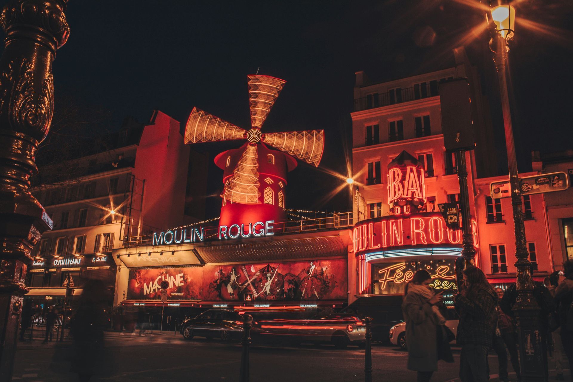 The Moulin Rouge building at night, with its iconic red windmill and neon signs brightly illuminated over a busy street.