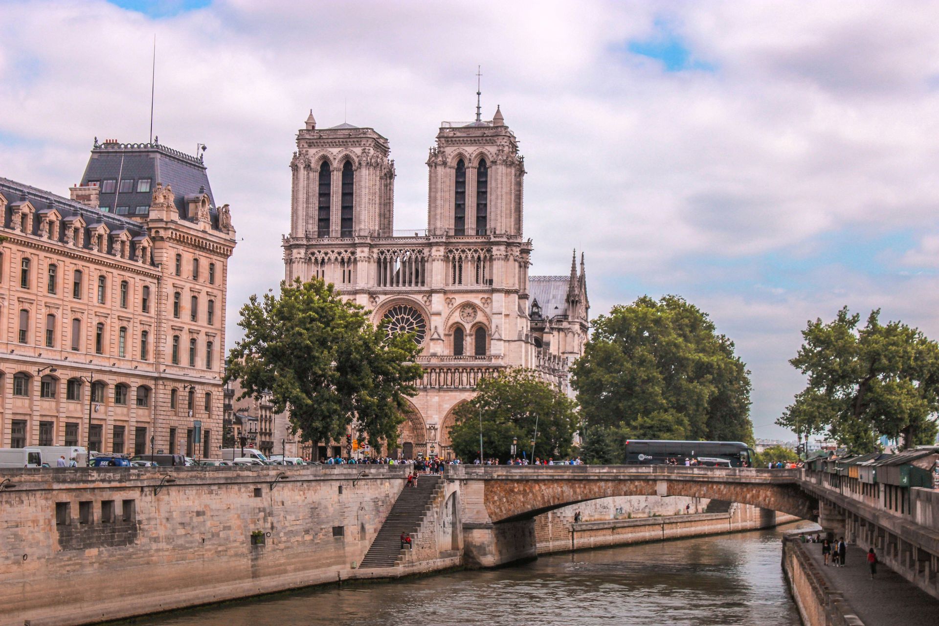 A large Gothic cathedral with twin towers stands behind a stone bridge crossing a river in a city under a cloudy sky.