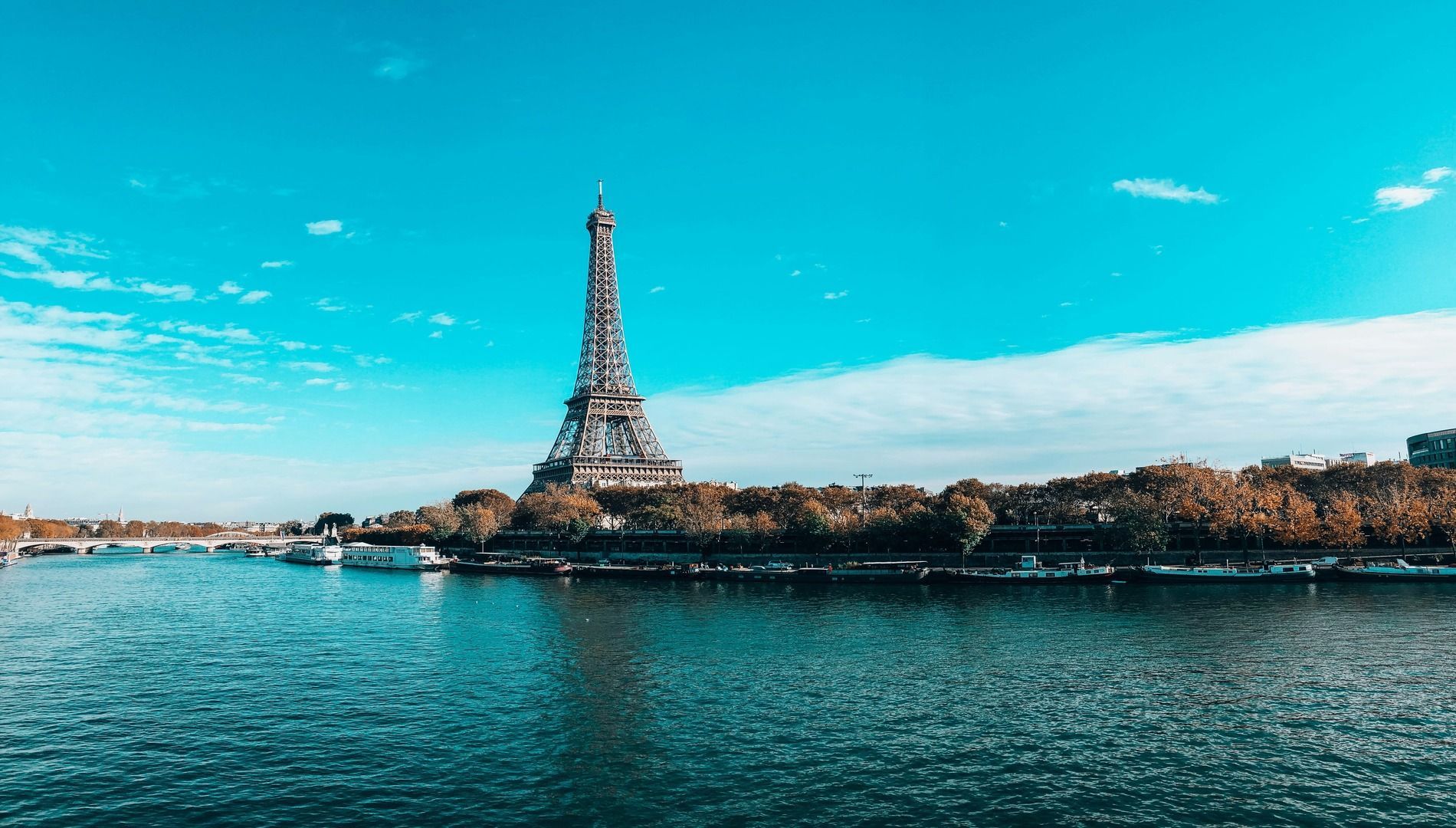 La Torre Eiffel se alza frente a un ancho río bordeado de árboles otoñales y barcos amarrados, bajo un cielo azul brillante.