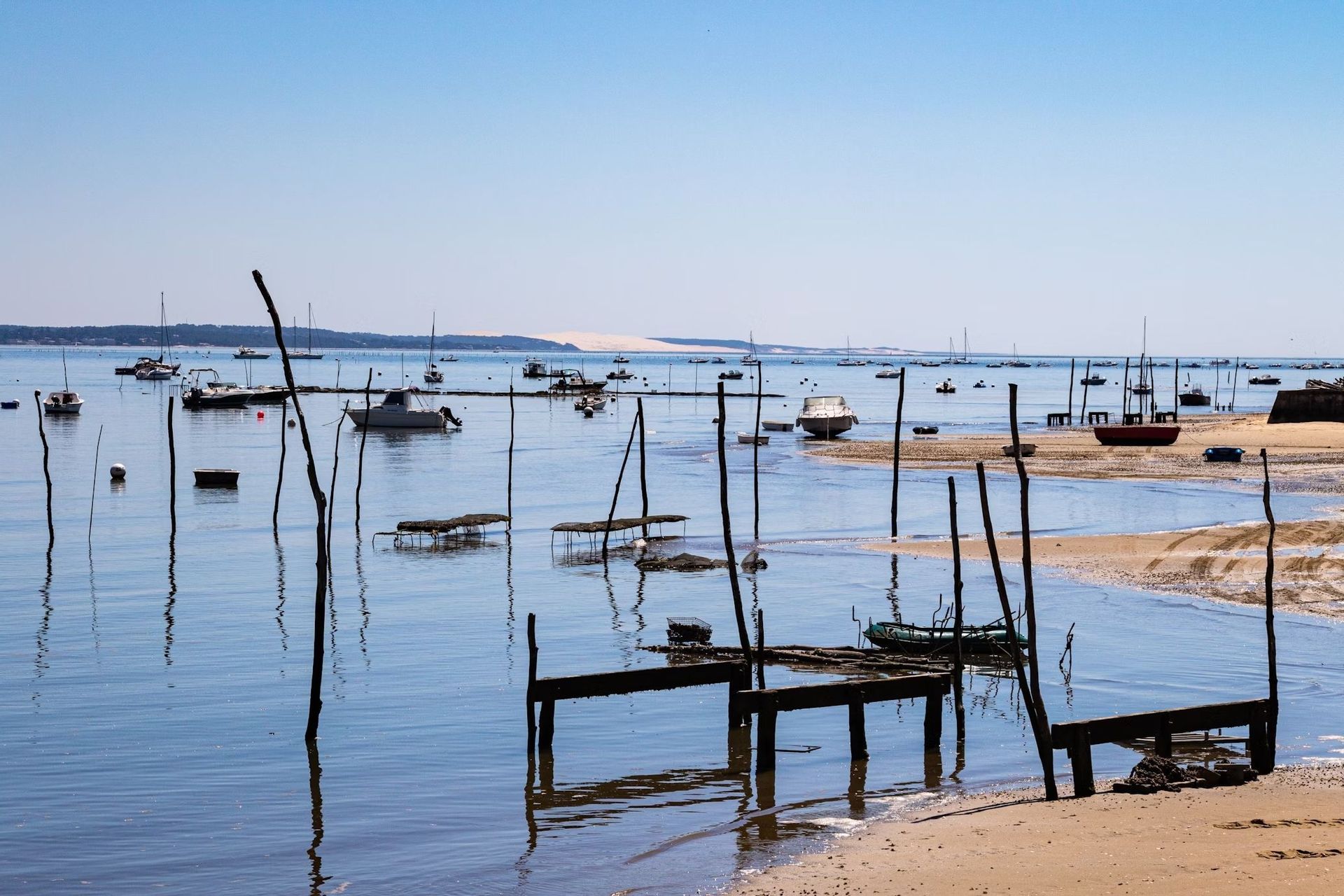 Petits bateaux amarrés dans une baie calme à marée basse, avec des piquets en bois sur la rive sablonneuse et une grande dune de sable à l'horizon lointain.