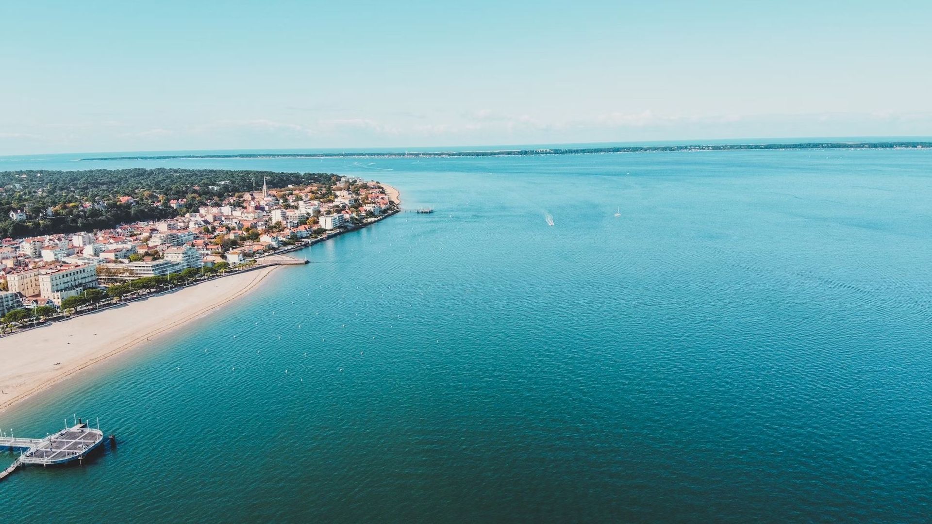 Vue aérienne d'une ville côtière avec une plage de sable bordant une mer bleue calme, avec une petite jetée au premier plan.