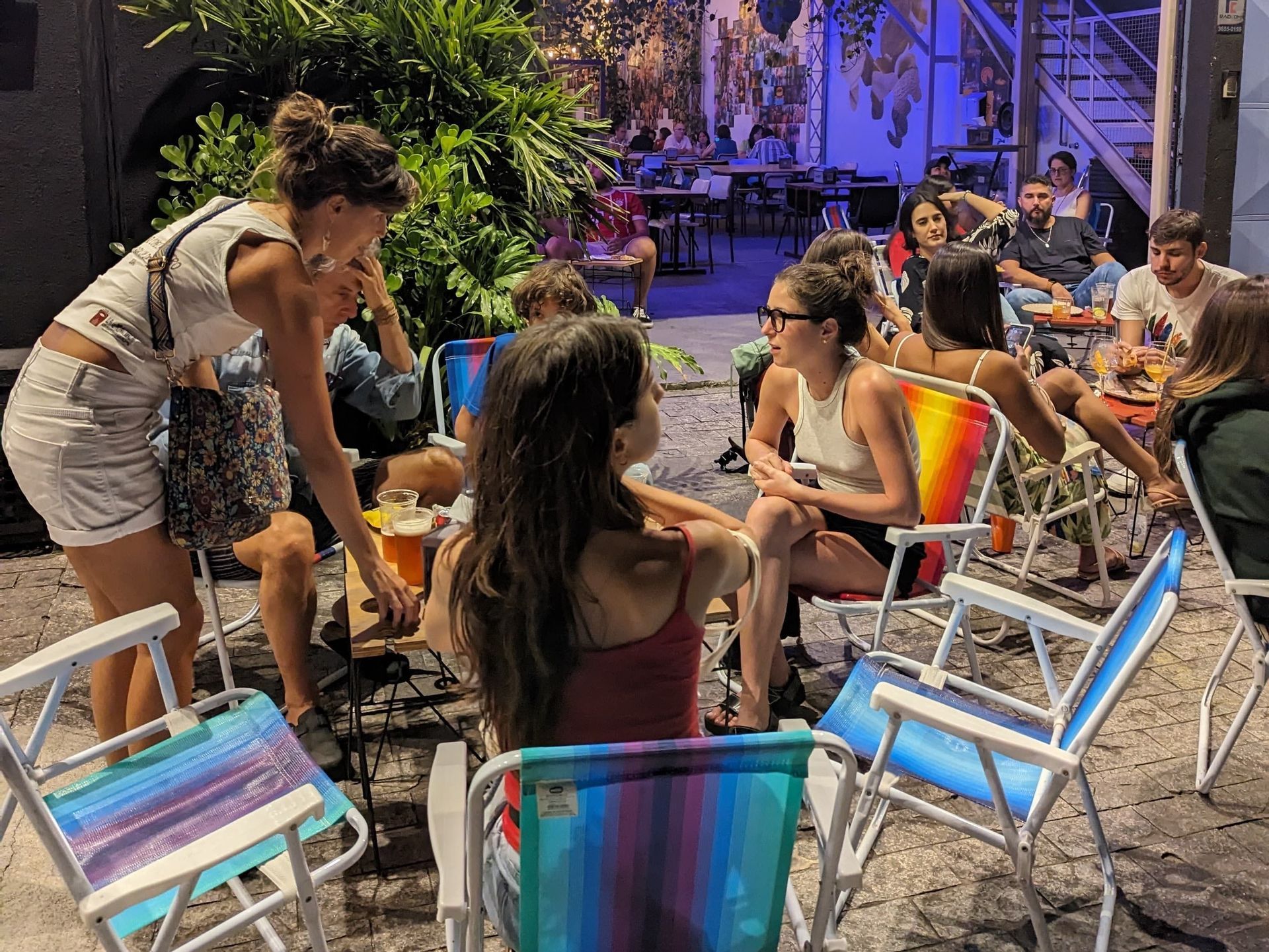 Un gruppo WeRoad si riunisce di notte in un cortile all'aperto, socializzando su sedie a sdraio colorate con drink.