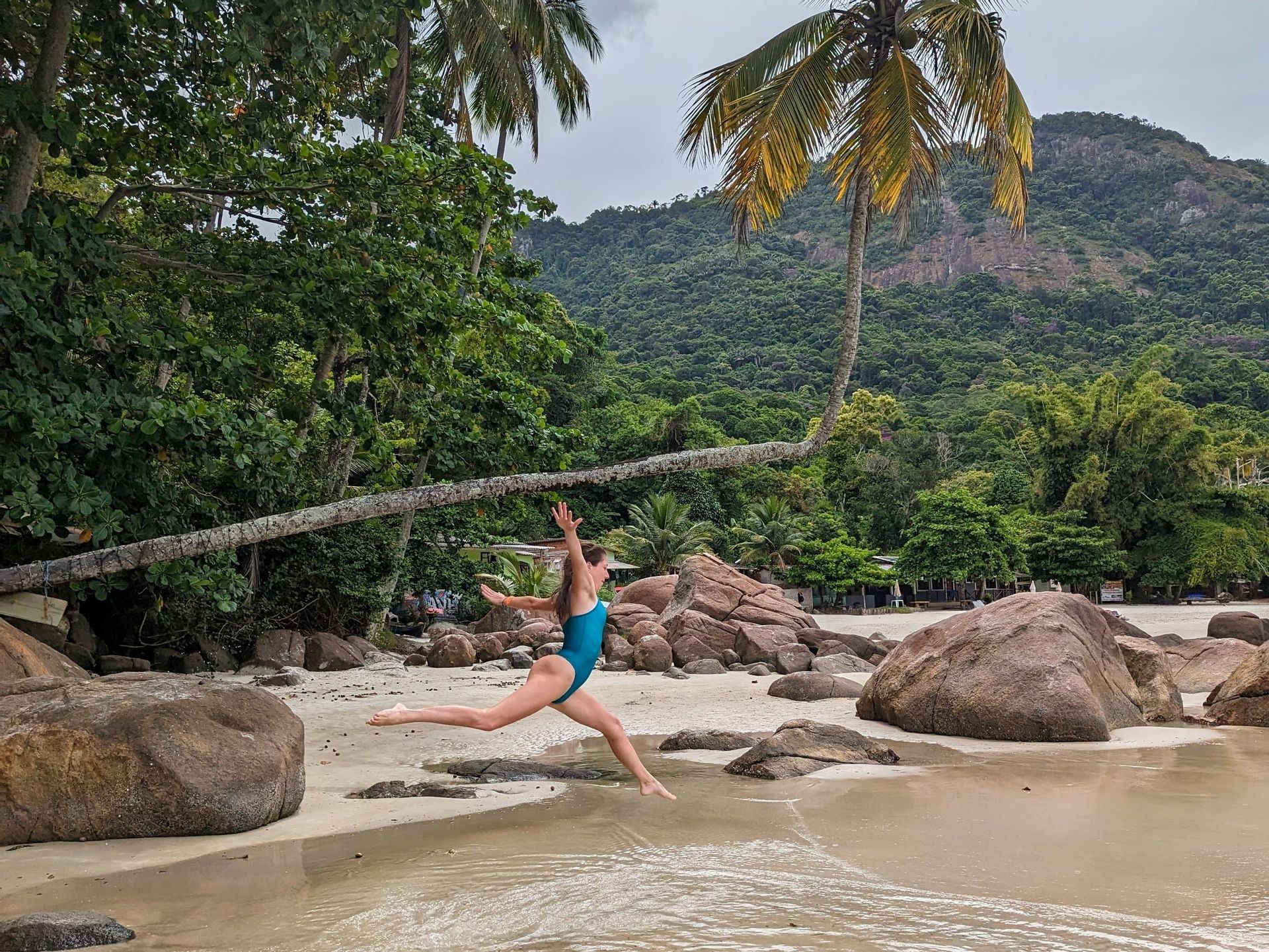 Una mujer en traje de baño azul salta en el aire en una playa de arena con grandes rocas, palmeras y una montaña boscosa de fondo.