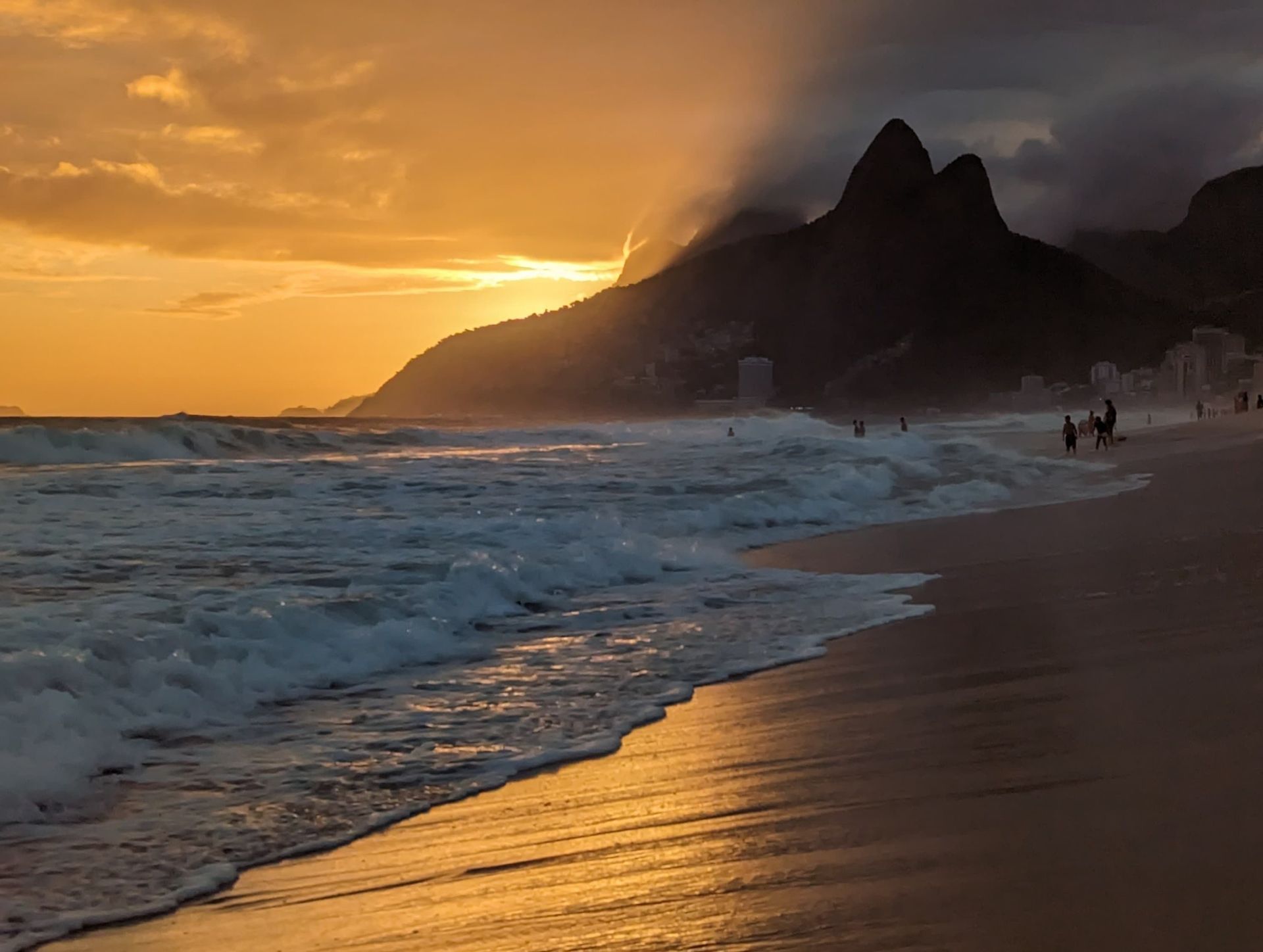 Ozeanwellen schlagen bei Sonnenuntergang an einem Sandstrand auf, wobei goldenes Licht auf dem Wasser reflektiert wird und sich dahinter silhouettierte Berge abzeichnen.