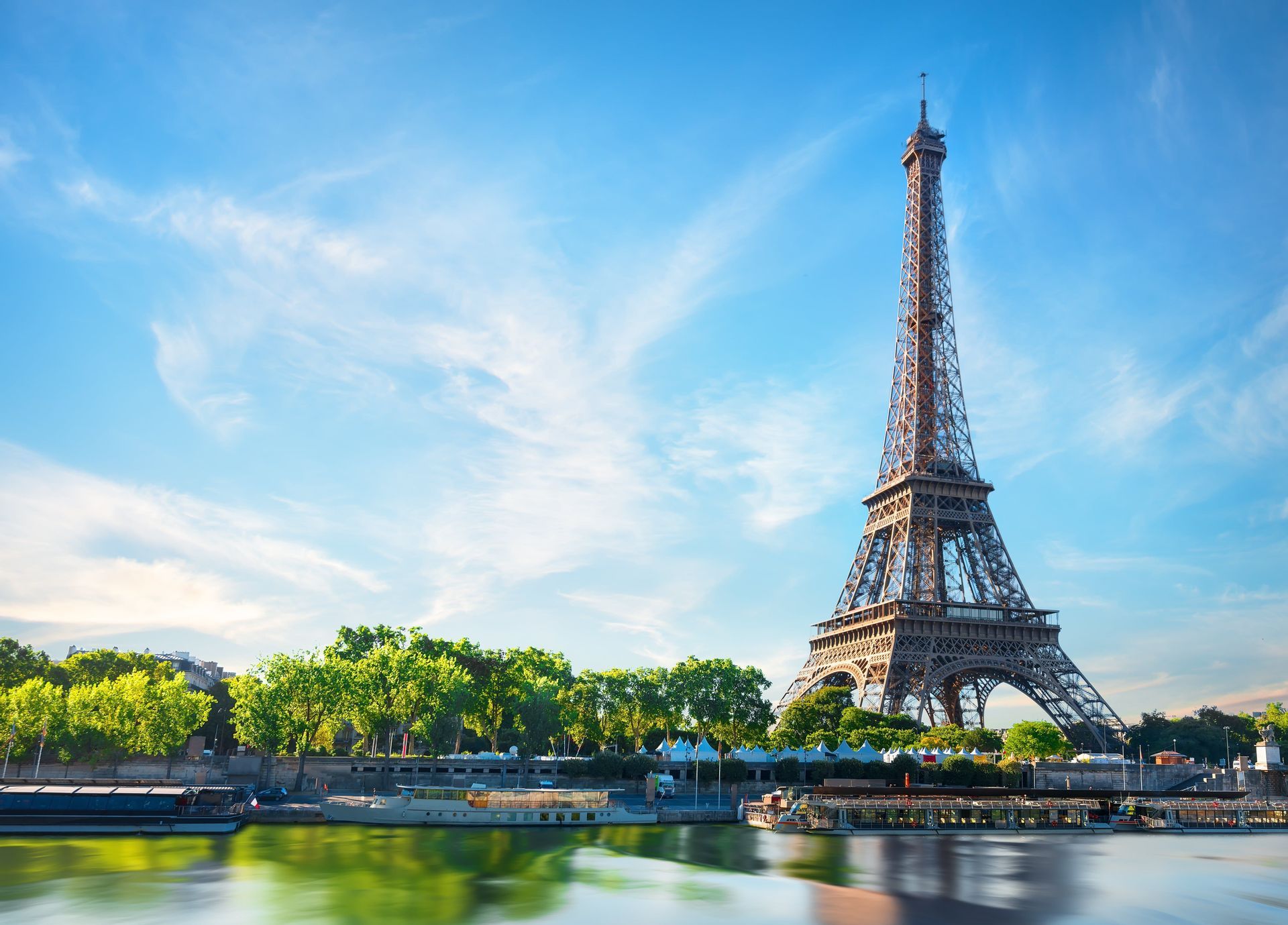La Torre Eiffel si erge sul fiume Senna con barche ormeggiate lungo la riva alberata sotto un cielo azzurro e limpido.
