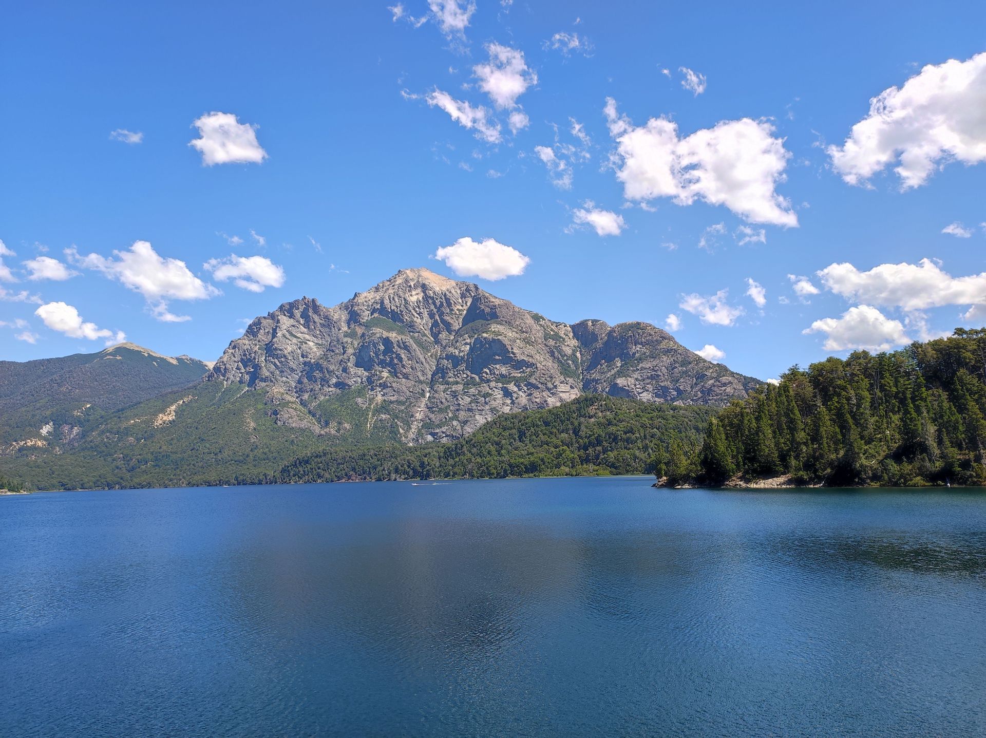 Una grande montagna rocciosa con pendii boscosi si erge sopra un lago blu calmo sotto un cielo parzialmente nuvoloso.