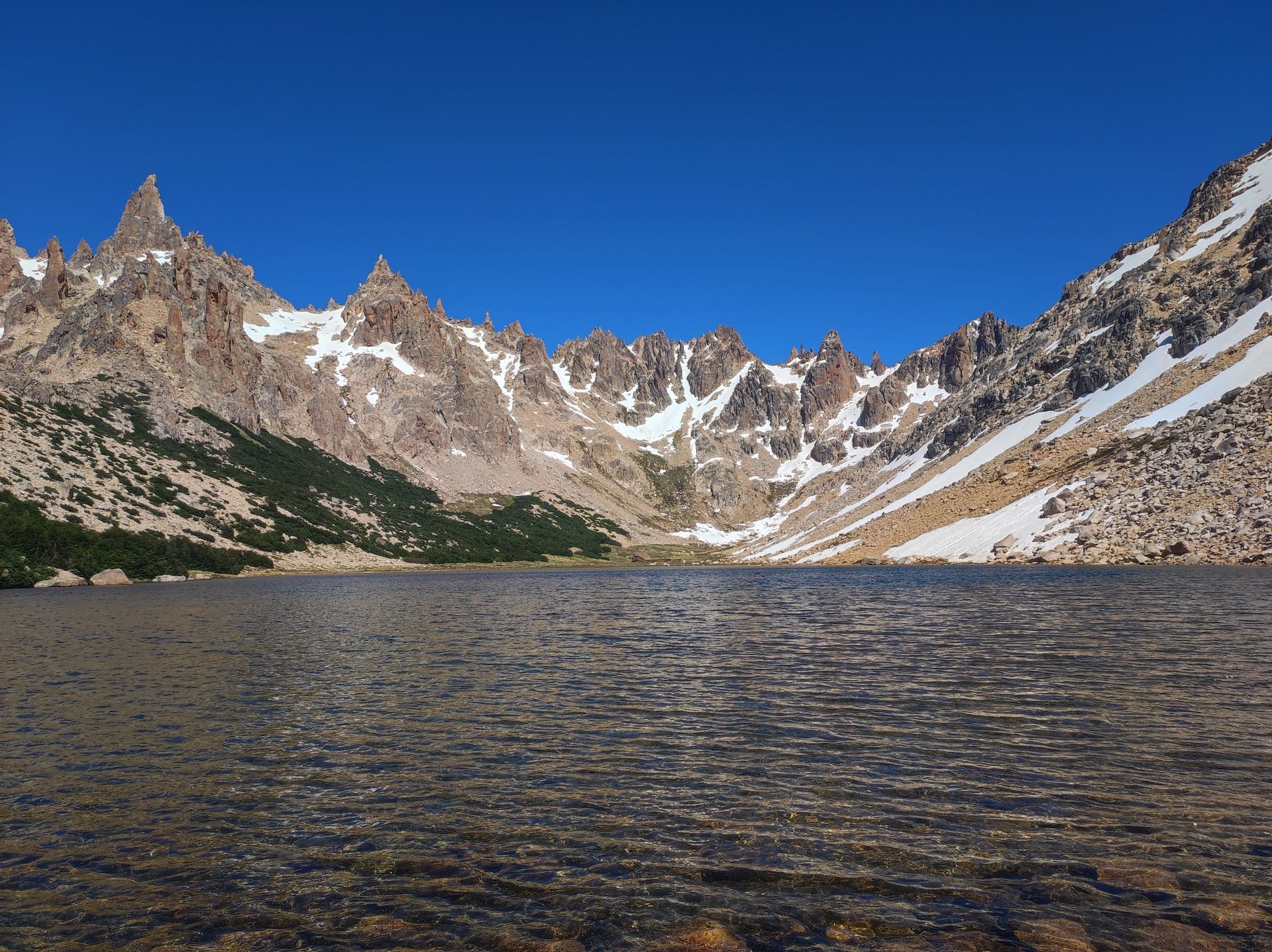 Un limpido lago di montagna riflette una catena montuosa frastagliata e innevata sotto un cielo azzurro senza nuvole.