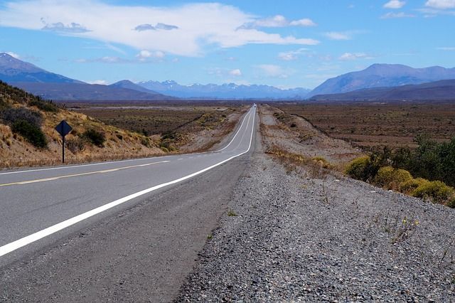 Una strada diritta e asfaltata si estende attraverso una vasta pianura arida verso una catena montuosa lontana sotto un cielo parzialmente nuvoloso.