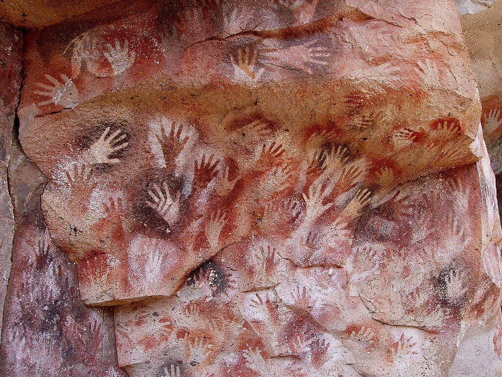 Antiche pitture rupestri con numerosi stencil di mani in pigmenti rossi, bianchi e scuri su una parete rocciosa grezza.