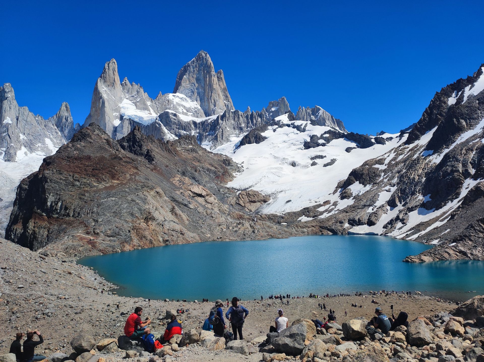 Un viaggio di gruppo WeRoad si rilassa sulla riva rocciosa di un lago alpino turchese, ai piedi di montagne frastagliate e innevate.