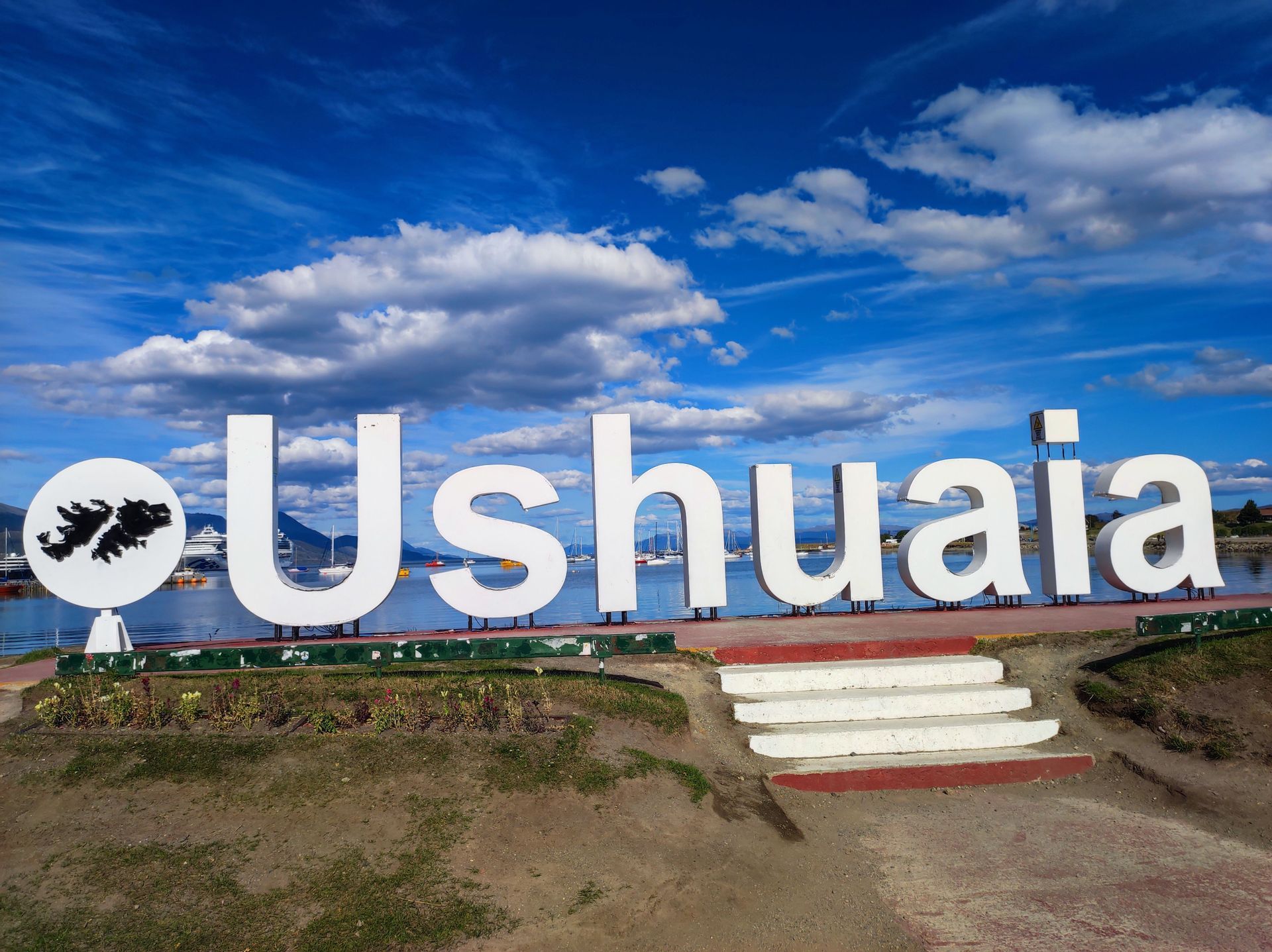Il grande cartello bianco 'Ushuaia' si erge di fronte a un porto con barche sotto un cielo blu parzialmente nuvoloso.